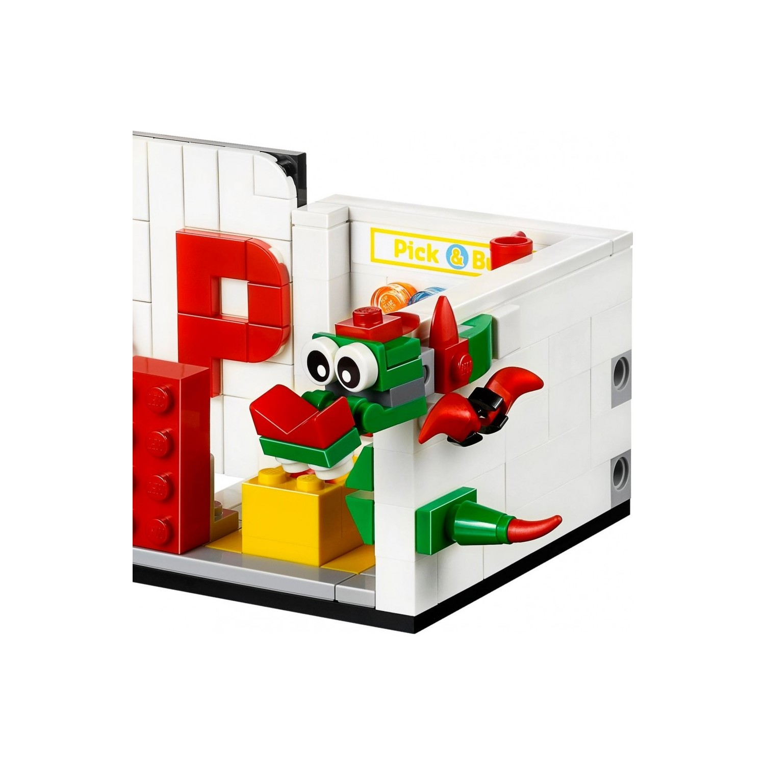 Ein grüner Lego-Drache mit roten Akzenten steht vor einer weißen Wand mit einem roten P.