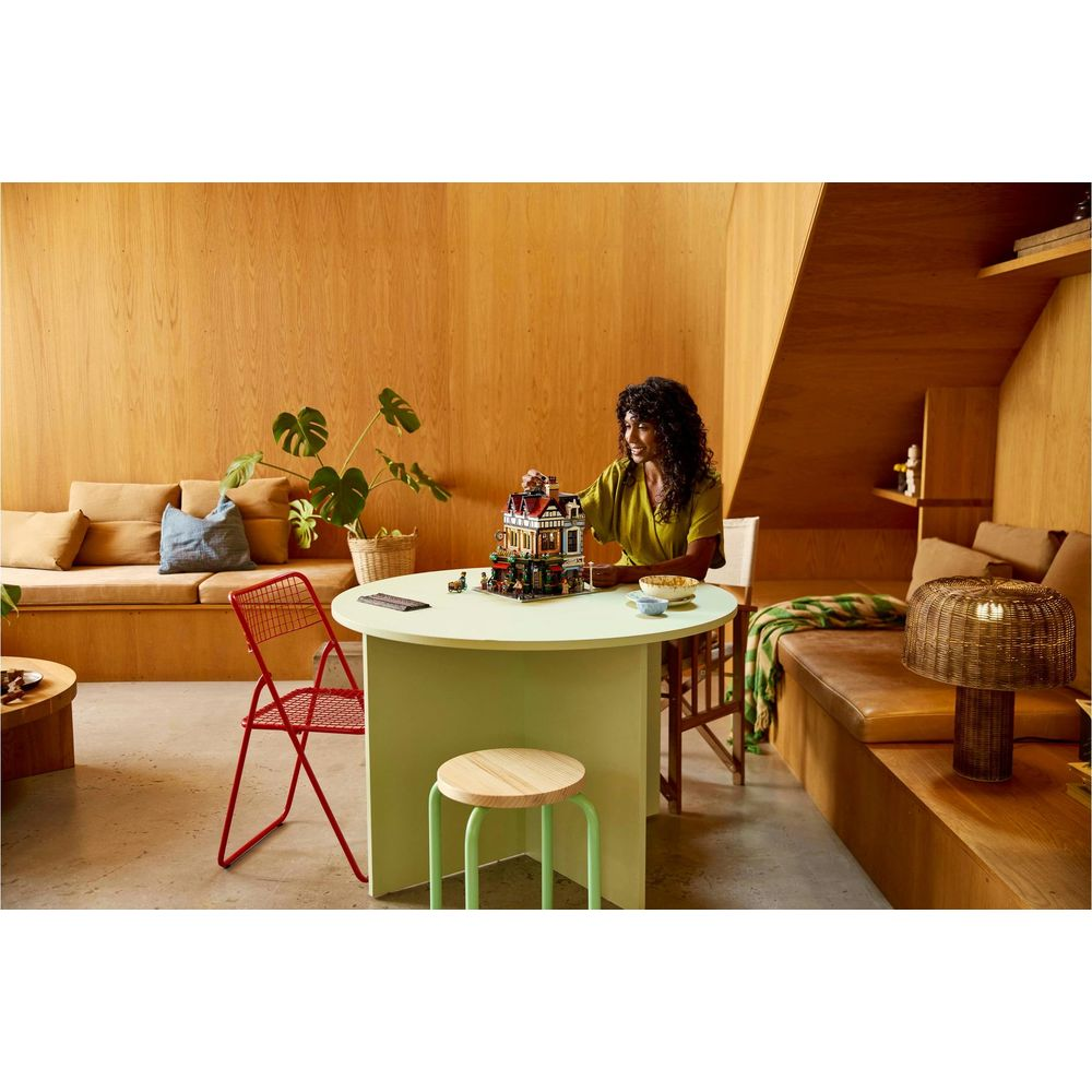 Kobieta siedzi przy stole, budując dom z klocków Lego. Drewniane ściany i sofy w tle.