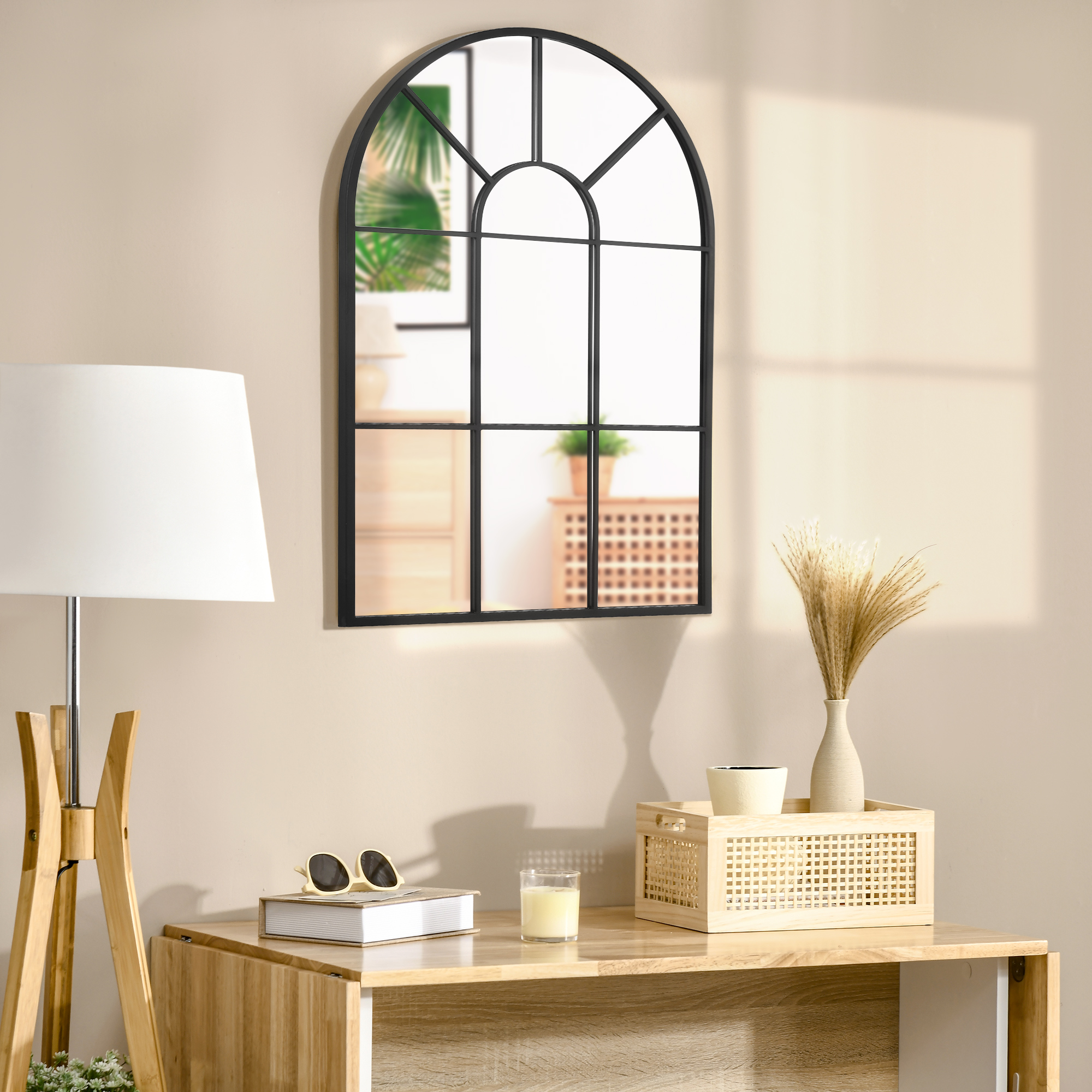 Un espejo de ventana arqueado sobre una mesa de madera con lámpara, libro, gafas de sol, vela y hierba seca.