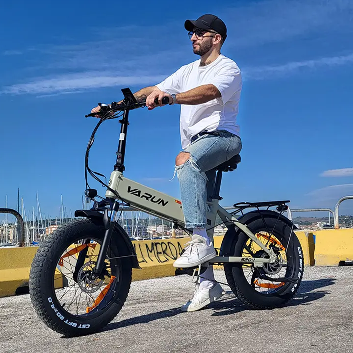 Mężczyzna na szarym rowerze elektrycznym z grubymi oponami, dżinsach, białej koszulce, czapce.