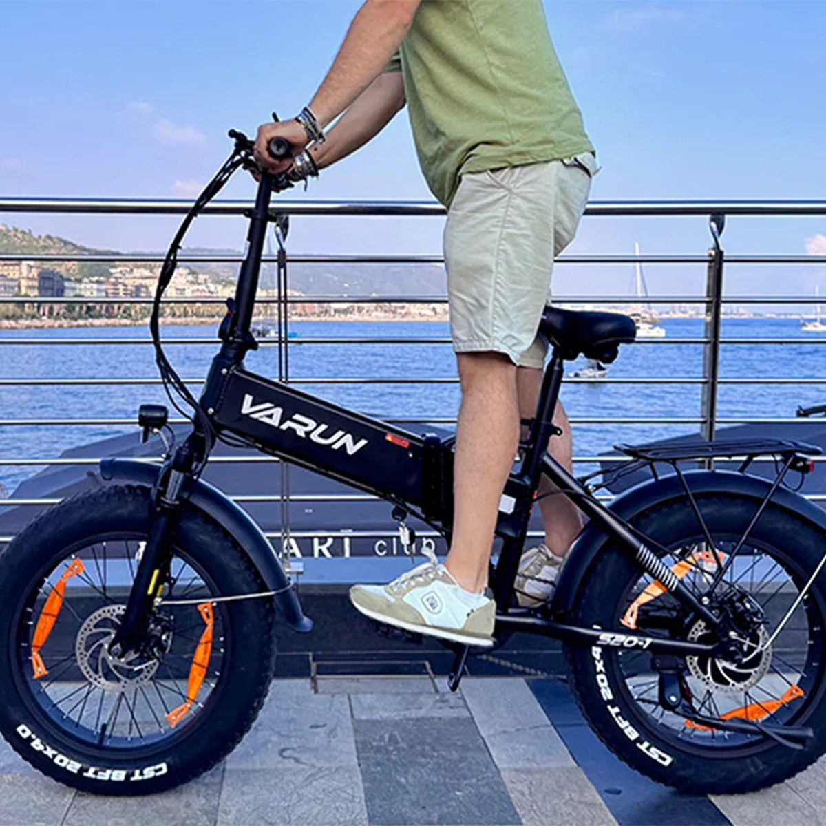 Mężczyzna na czarnym rowerze elektrycznym z szerokimi oponami nad wodą, miasto i morze w tle.