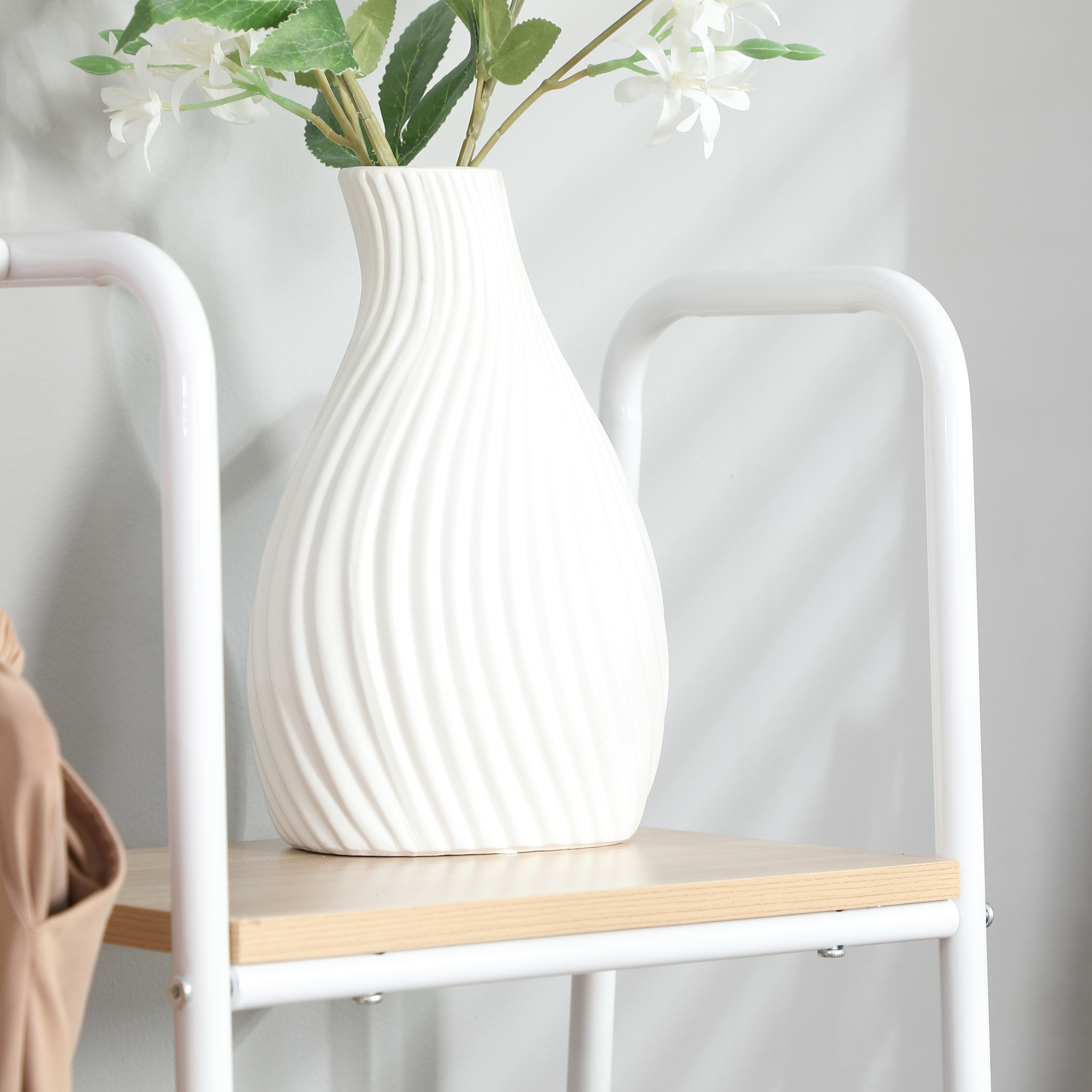 Un jarrón blanco con flores en una estantería. Ropa beige a la izquierda.