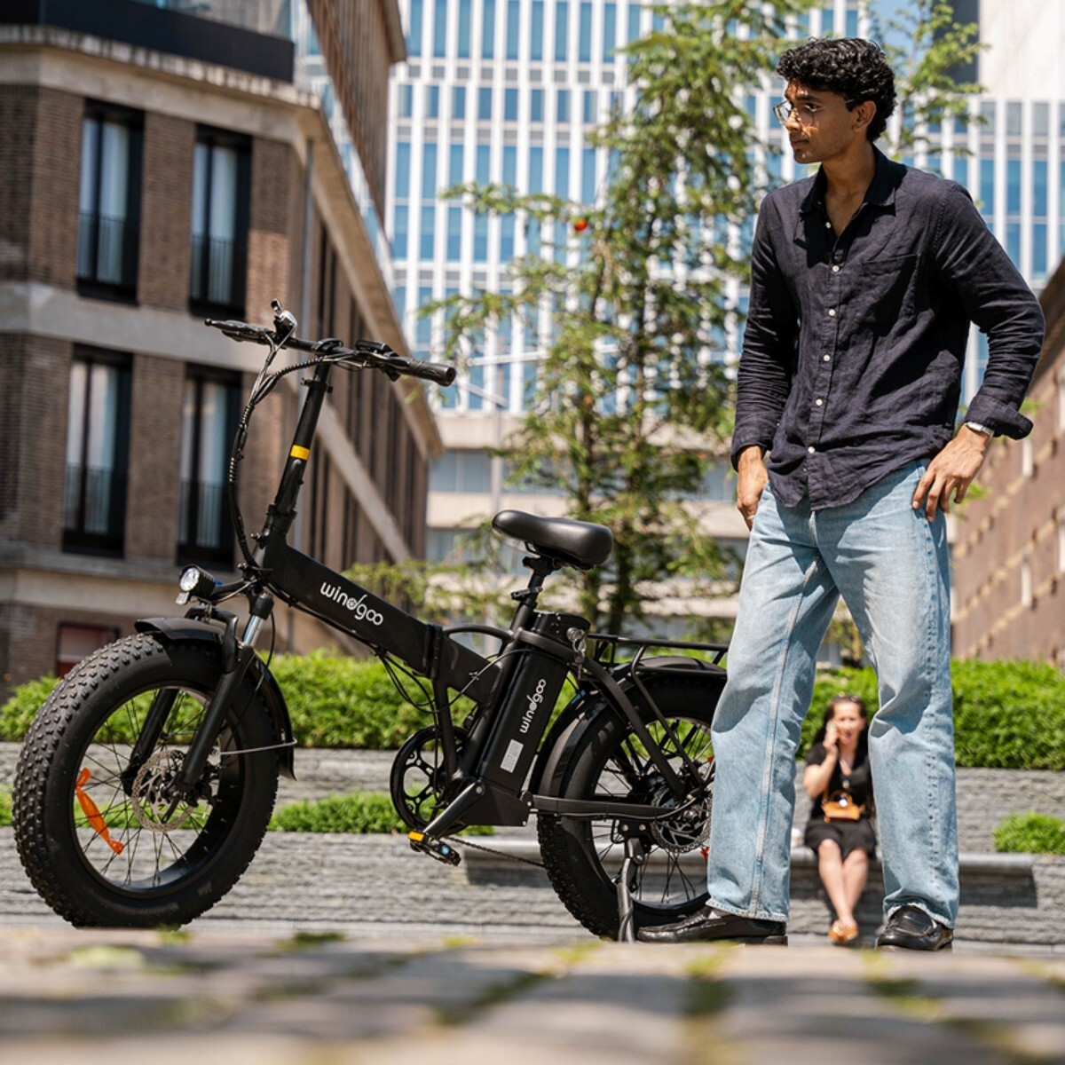 Mężczyzna w dżinsach i ciemnej koszuli stoi obok czarnego roweru elektrycznego. Ludzie siedzą w tle.