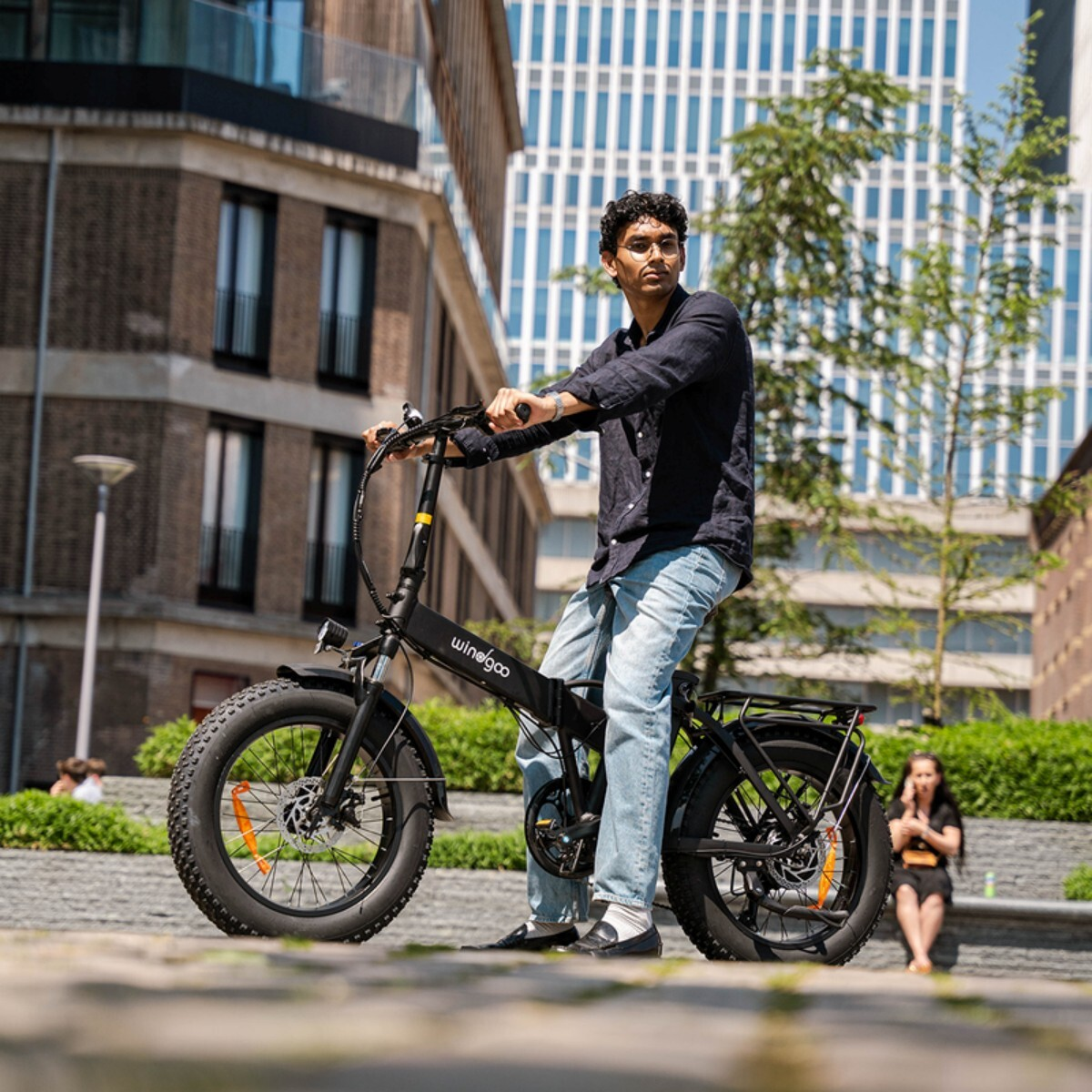 Mężczyzna na czarnym rowerze elektrycznym z szerokimi oponami przed nowoczesnymi budynkami.