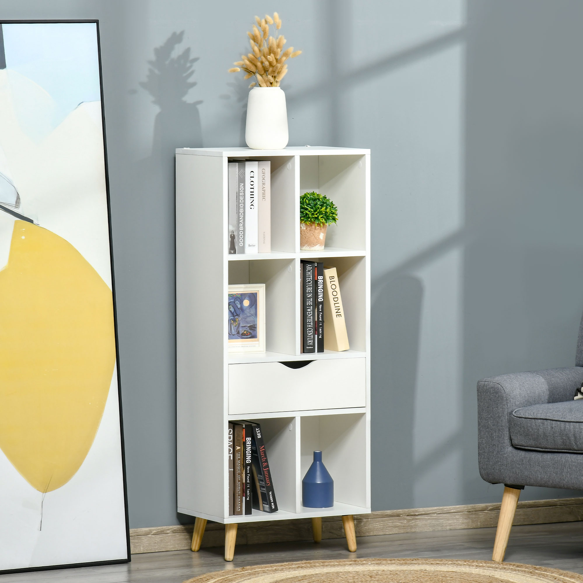 Una estantería blanca con libros, jarrón, maceta y cuadro junto a un sillón.