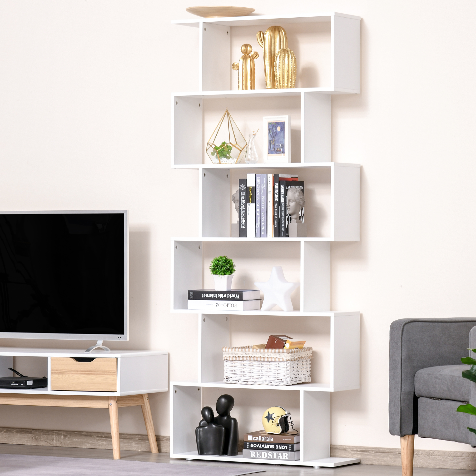 Una estantería blanca en zigzag muestra decoración, libros y una cesta. A la izquierda hay un televisor y un soporte.