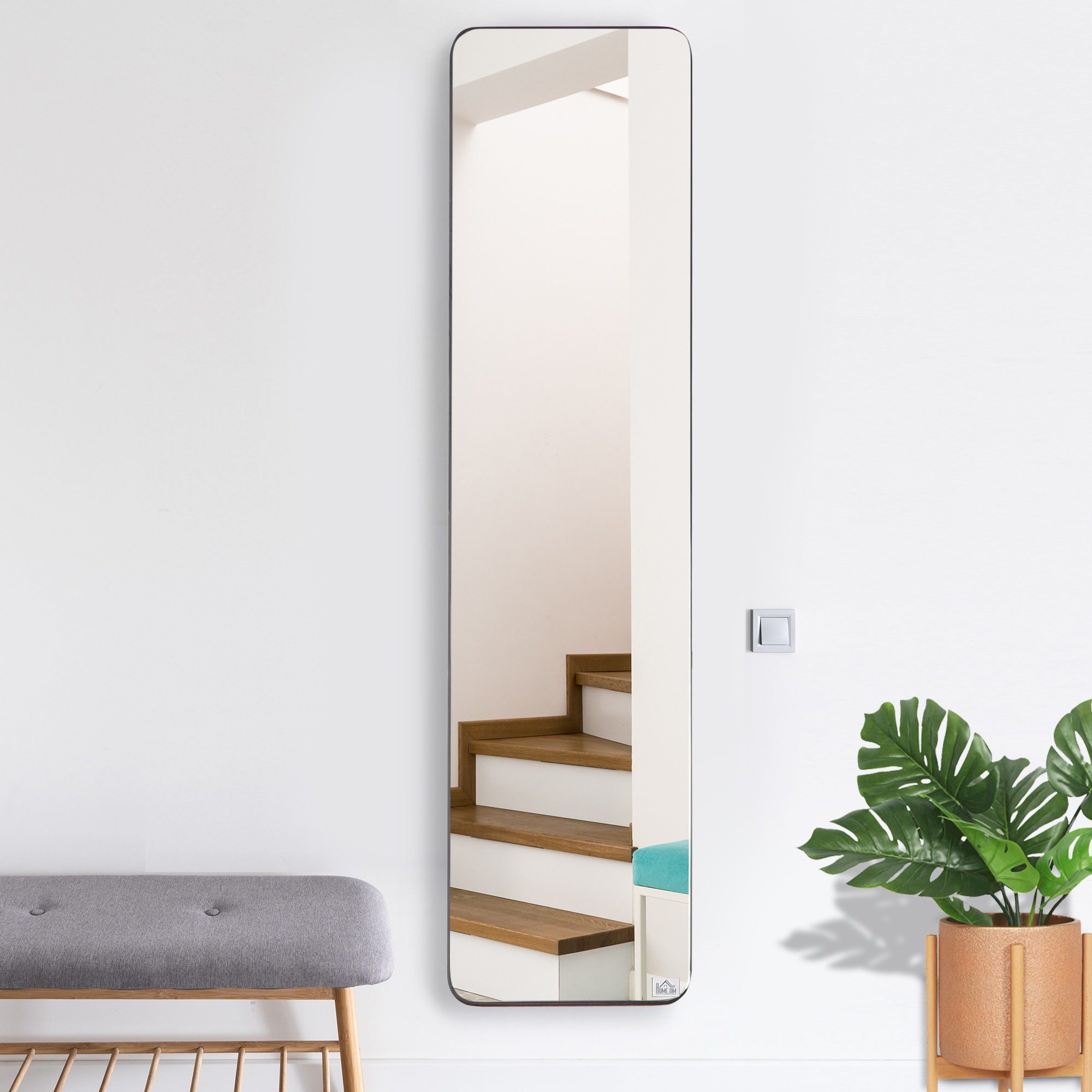 Un espejo alto refleja una escalera de madera. Se ve un banco gris y una planta.