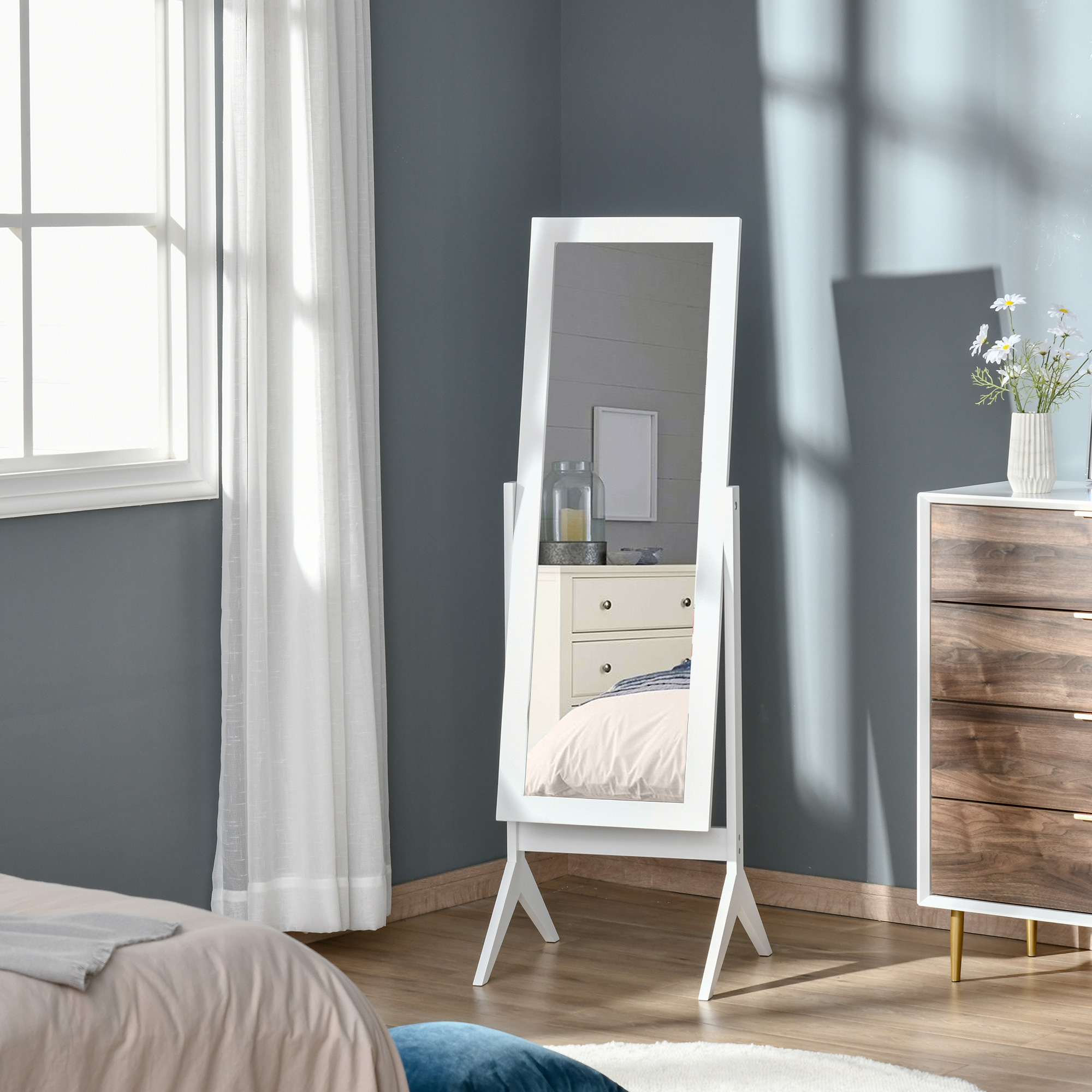 Un espejo de pie blanco está junto a una cómoda con flores. Una ventana con cortinas está a la izquierda.