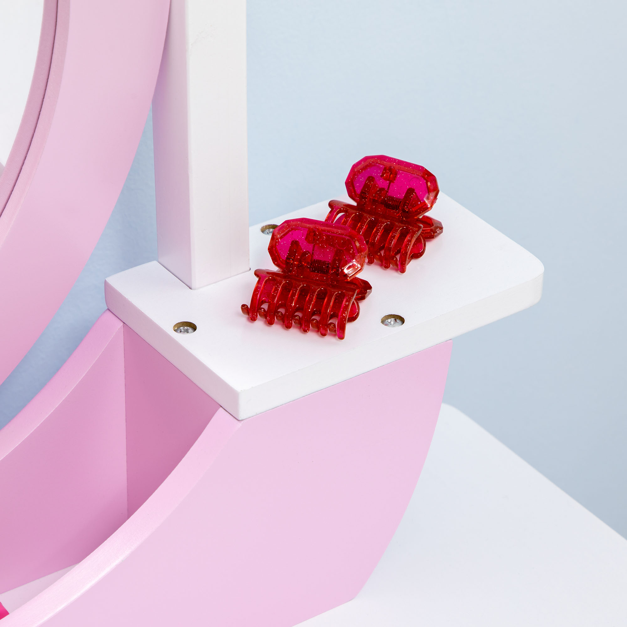 Dos pinzas para el pelo rojas y brillantes descansan en un estante blanco, parte de un tocador rosa.