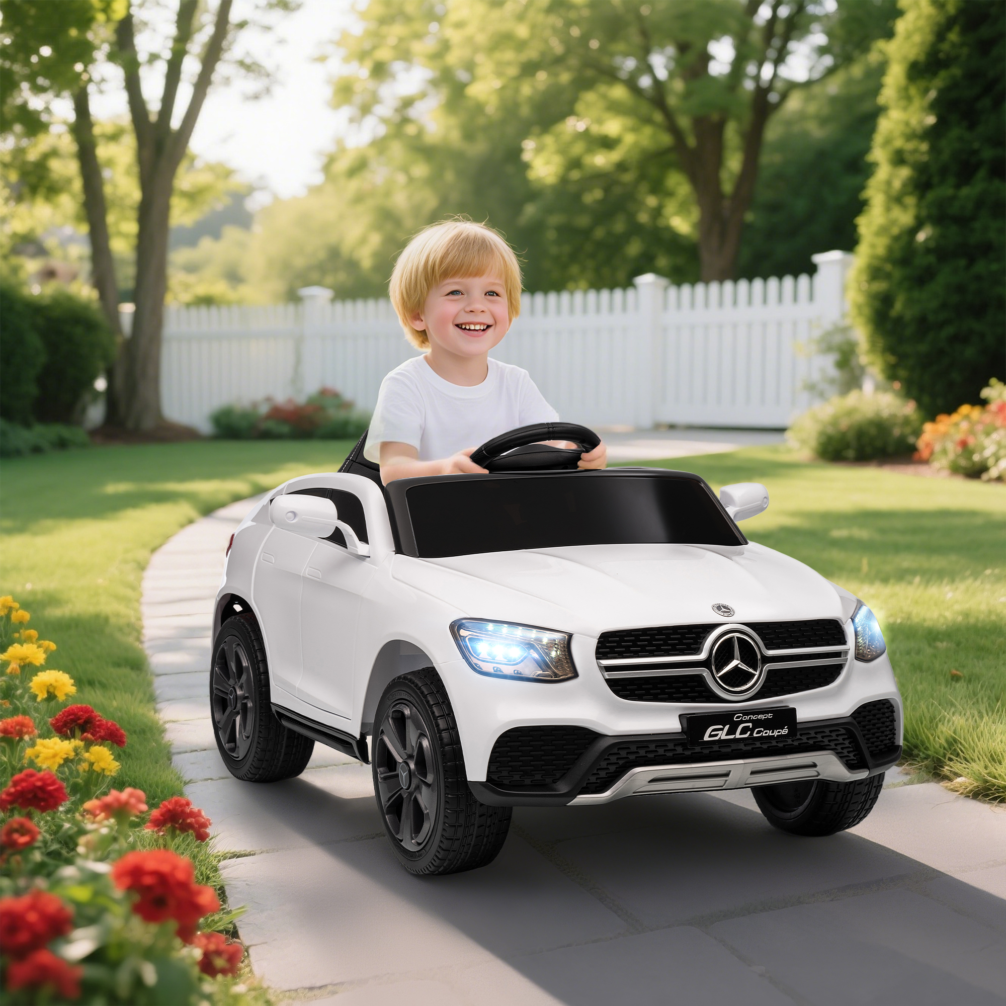 Un niño conduce un coche eléctrico blanco en un jardín con flores y una valla.