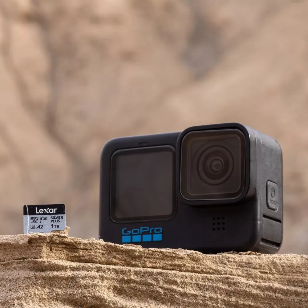 Una fotocamera GoPro nera e una scheda SD Lexar riposano su terreno sabbioso.