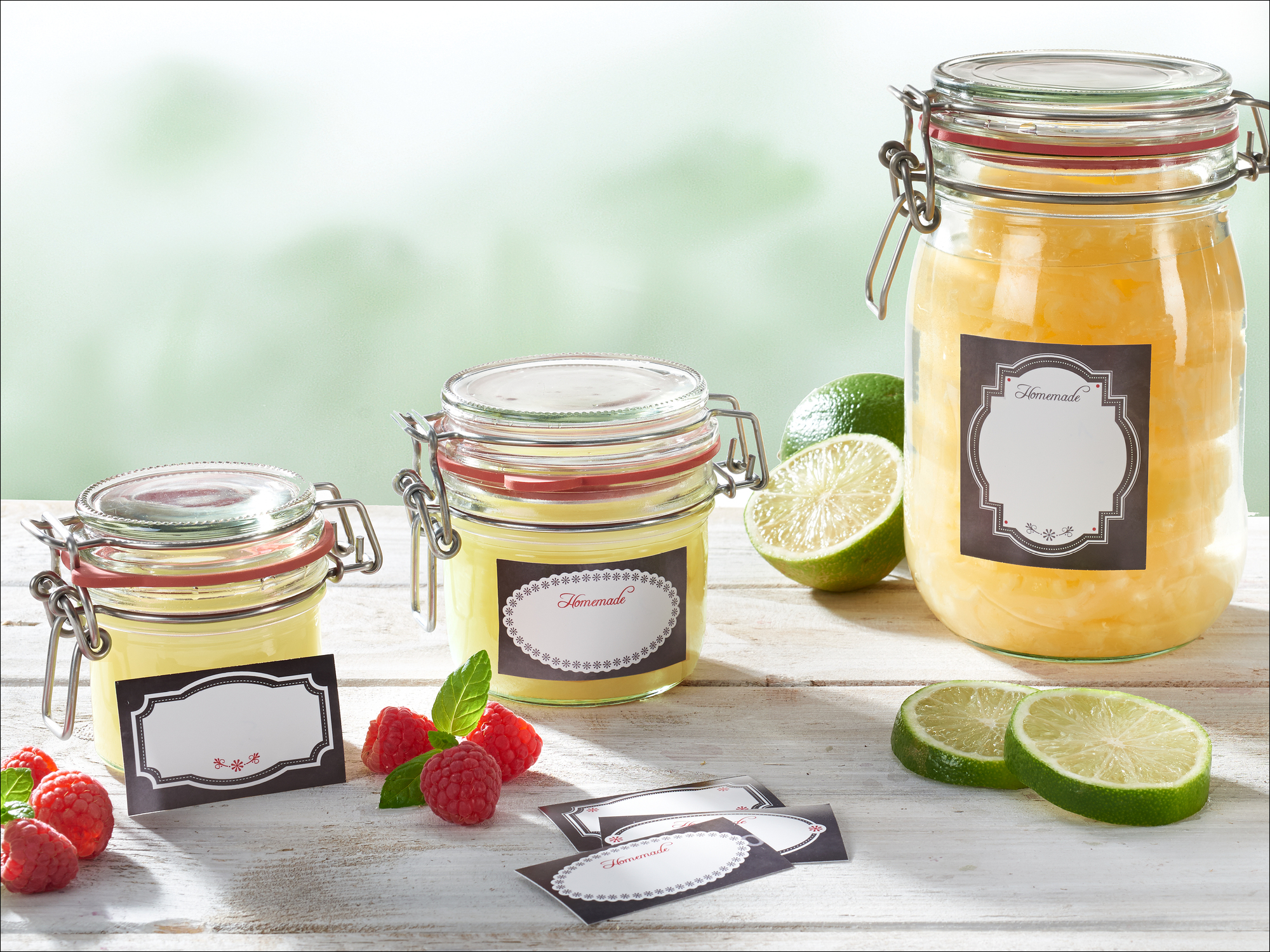 Drei Gläser mit gelbem Inhalt, Limetten, Himbeeren und Etiketten auf Holztisch.