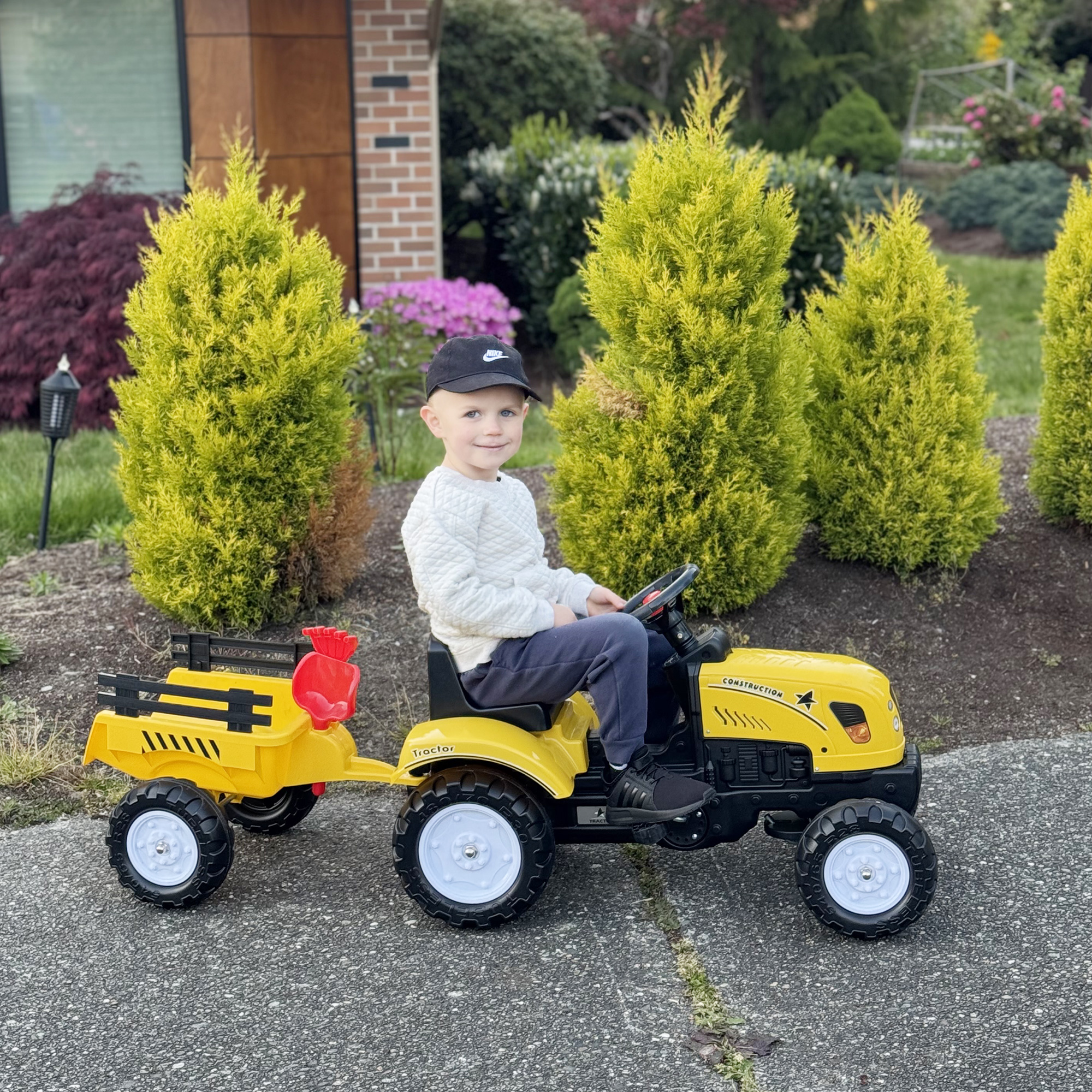 Un niño pequeño con suéter blanco y pantalones azules monta un tractor de juguete amarillo con remolque.