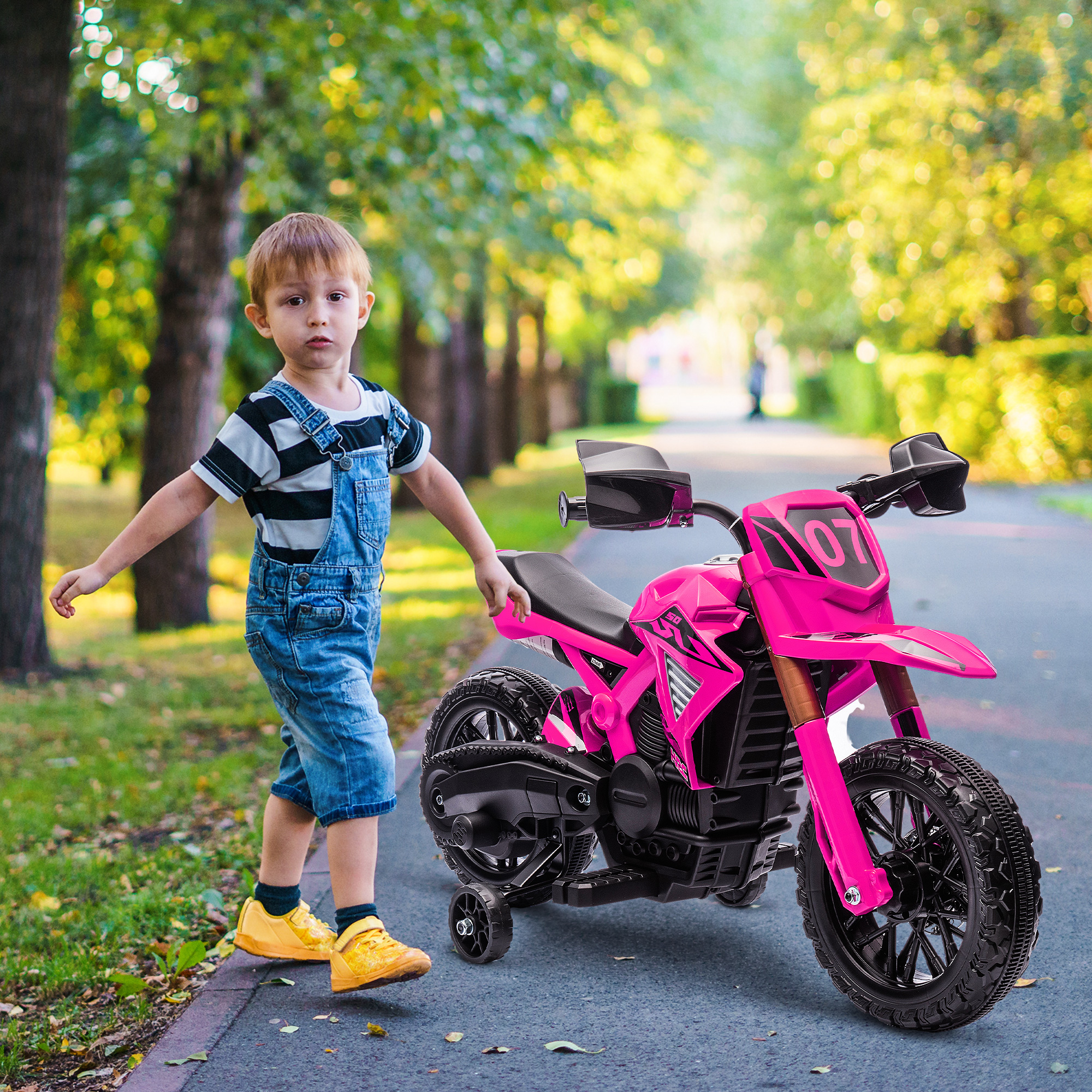 Un niño pequeño con peto está junto a una motocicleta de juguete rosa en un parque.