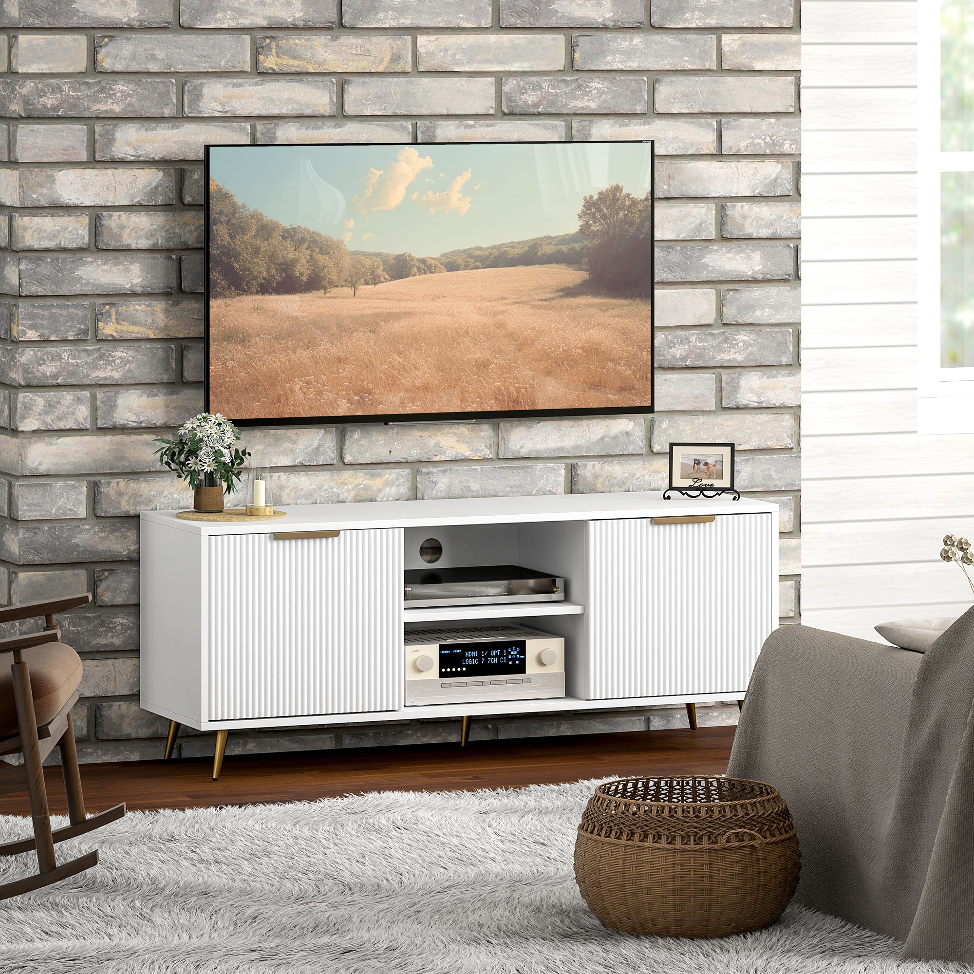 Un mueble de TV blanco con patas doradas frente a una pared de ladrillos. Un televisor muestra un paisaje.