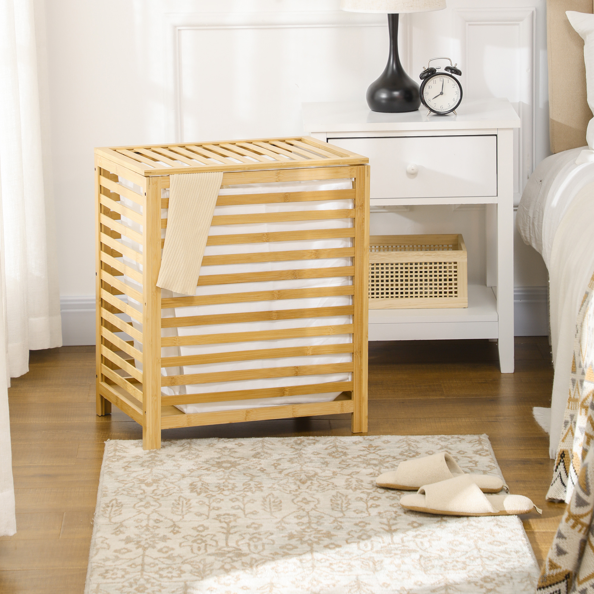 Una cesta de lavandería de madera junto a una mesita de noche con lámpara y despertador.