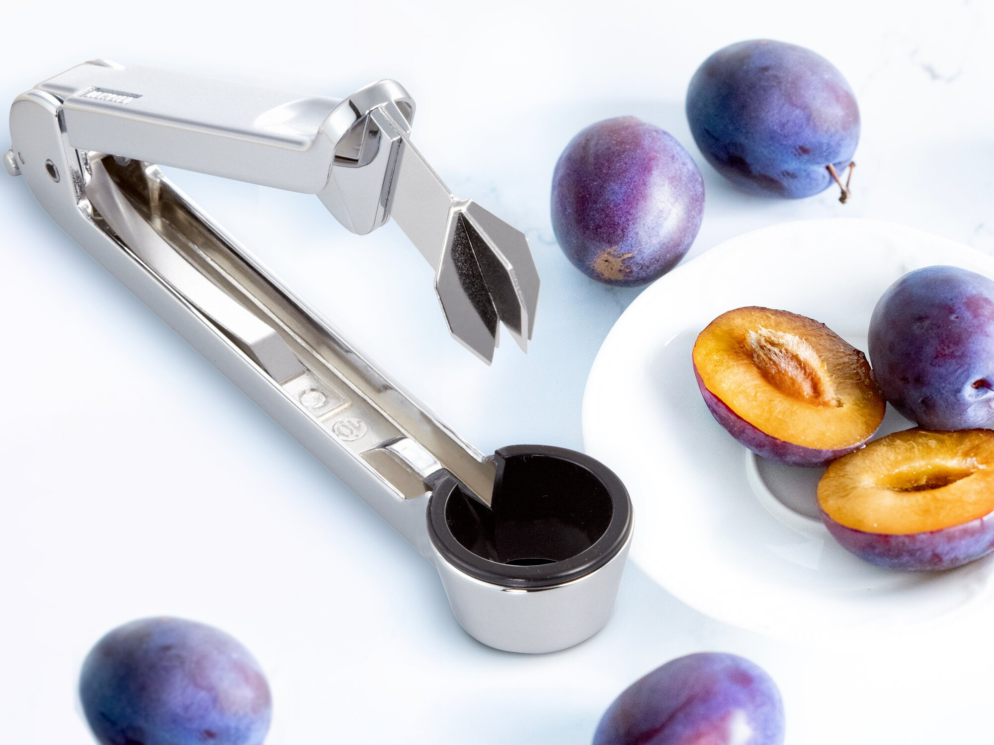 Ein silbernes Obstentkerngerät neben ganzen und halbierten Pflaumen auf weißem Hintergrund.