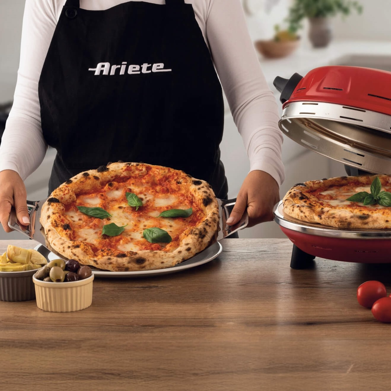 Person in schwarzer Schürze hält eine Pizza neben einem roten Pizzaofen.