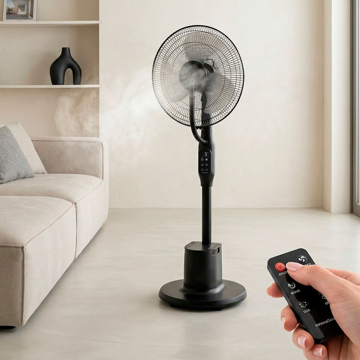 Un ventilatore da terra nero con nebulizzatore accanto a un divano. Una mano tiene un telecomando.