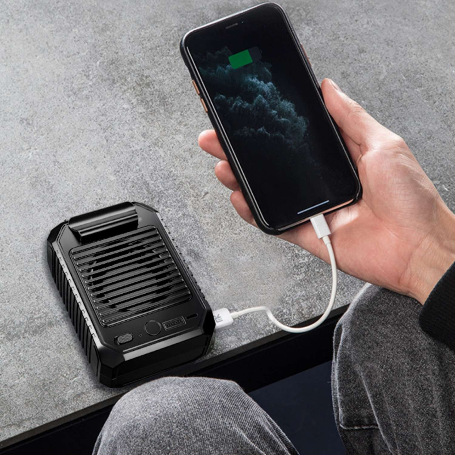 Ein schwarzer tragbarer Lüfter lädt ein Smartphone, verbunden durch ein weißes Kabel auf grauer Oberfläche.