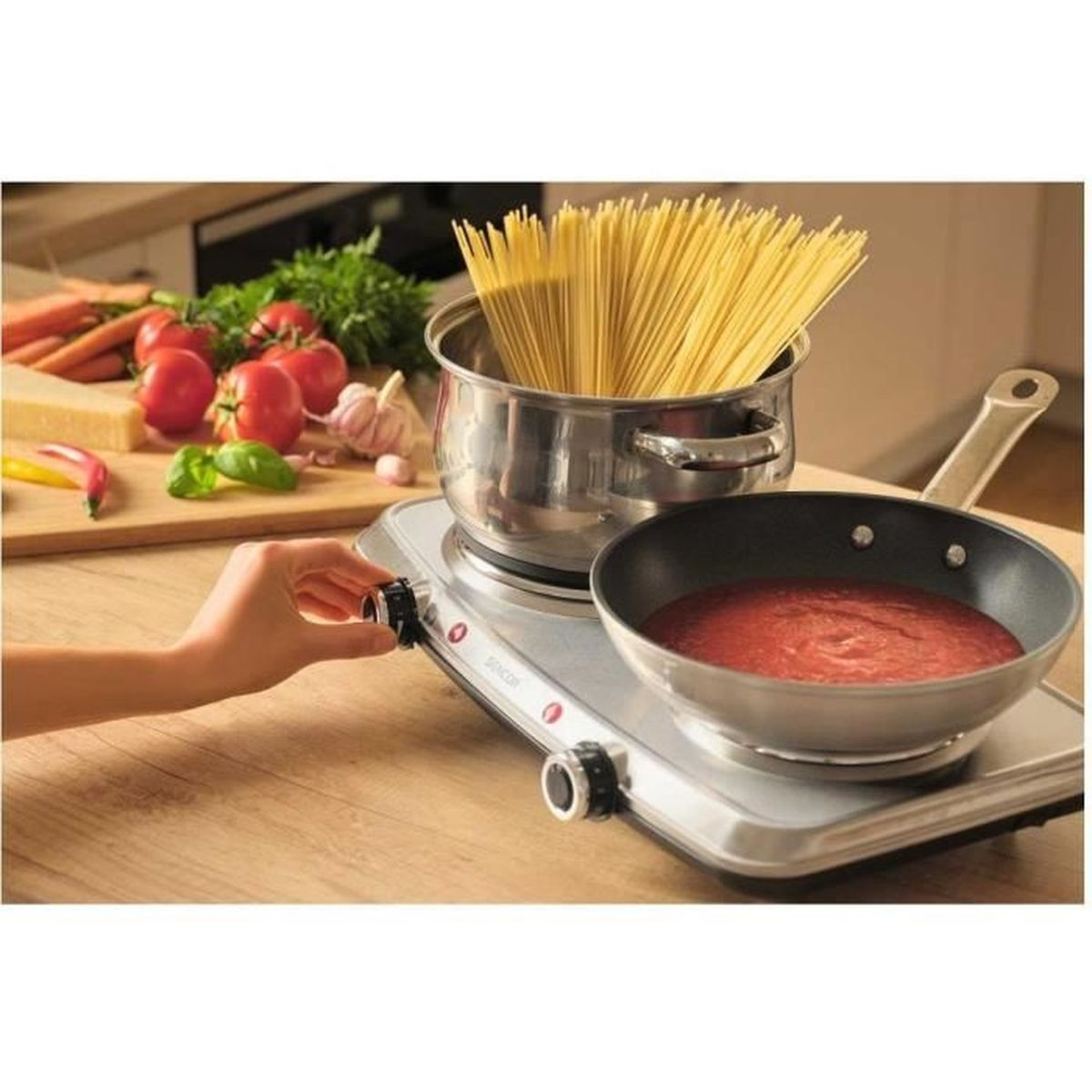 Una mano gira una manopola su un piano cottura doppio. C'è una pentola di spaghetti e una padella di sugo.