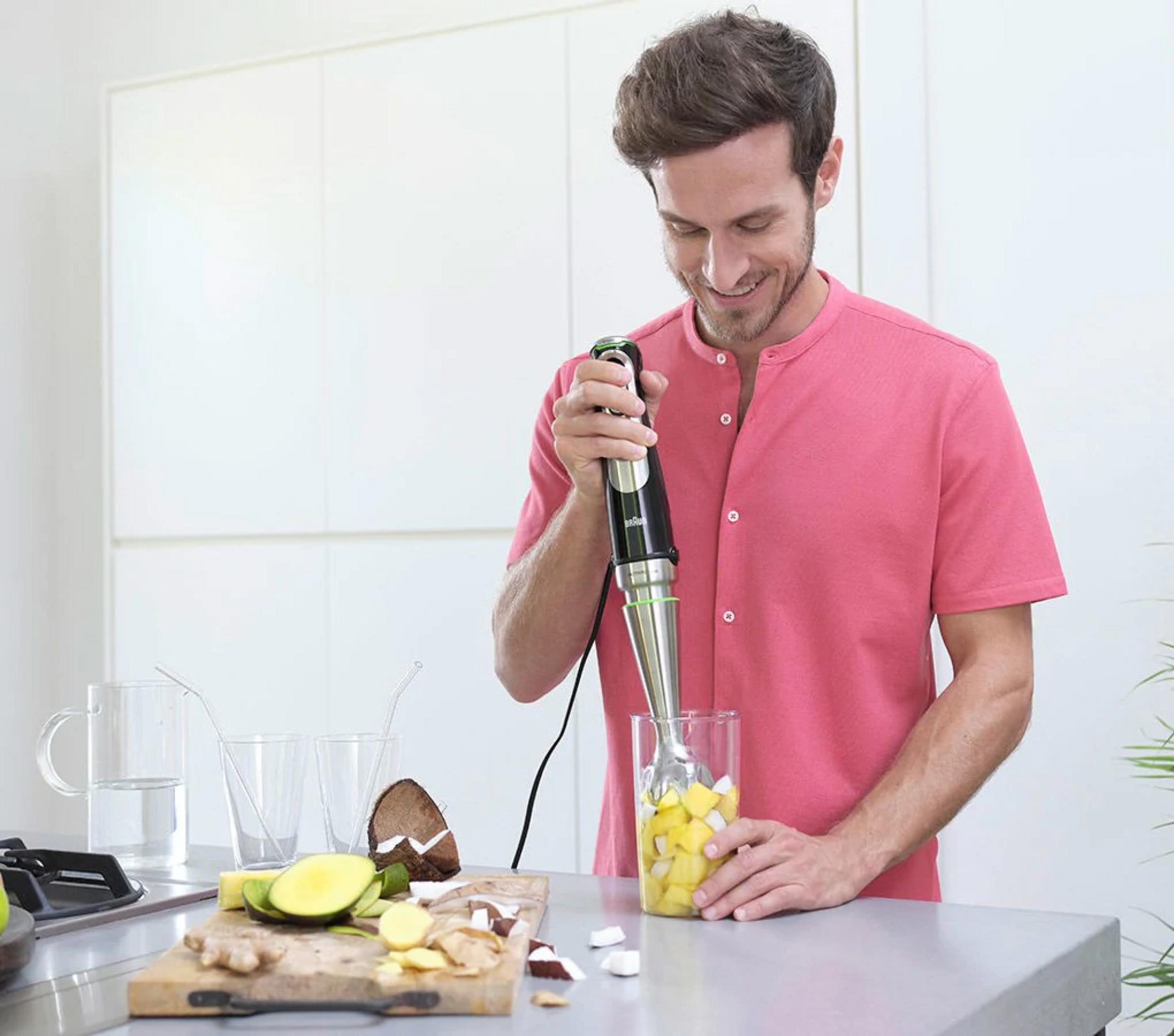 Een man mixt fruit in een glas met een staafmixer in een moderne keuken.