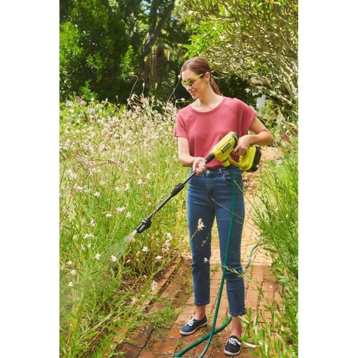 Frau wäscht im Garten mit einem Hochdruckreiniger Blumen und Pflanzen auf einem Ziegelweg.