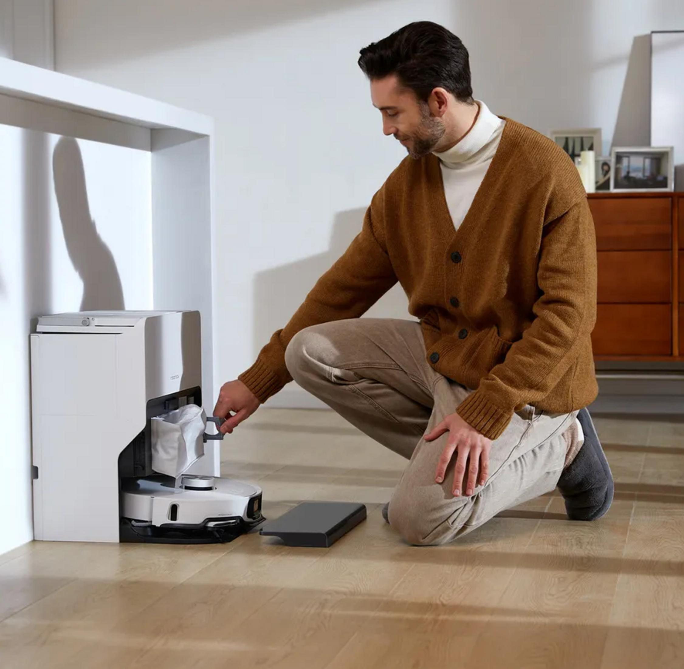 Mężczyzna w brązowym swetrze klęczy obok robota sprzątającego i jego stacji.