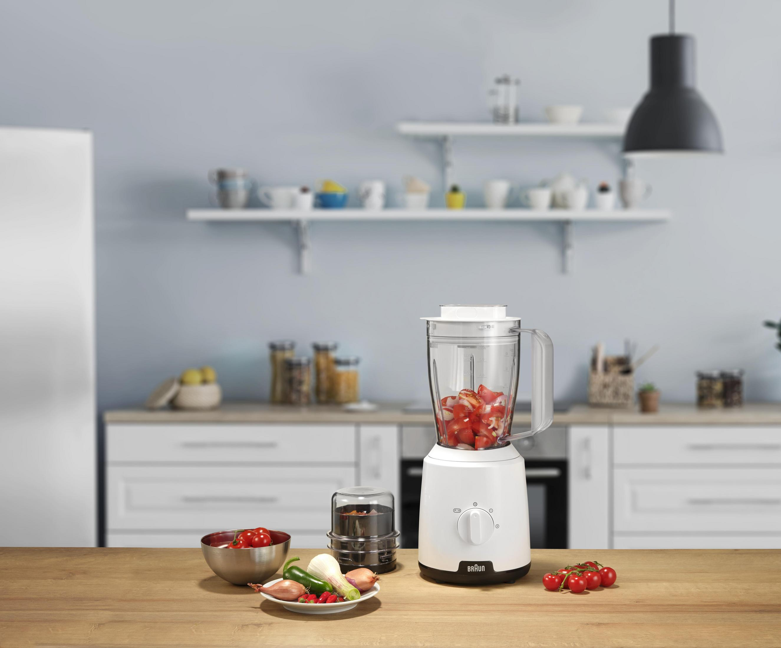 Een witte blender met tomaten en uien, ernaast groenten en kerstomaatjes.