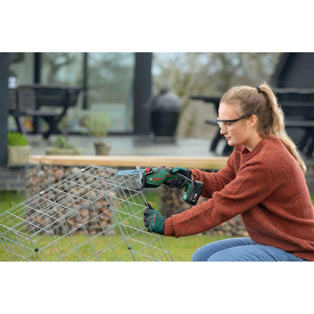 Frau mit Schutzbrille schneidet mit Säge Drahtgitter im Garten. Draußen.