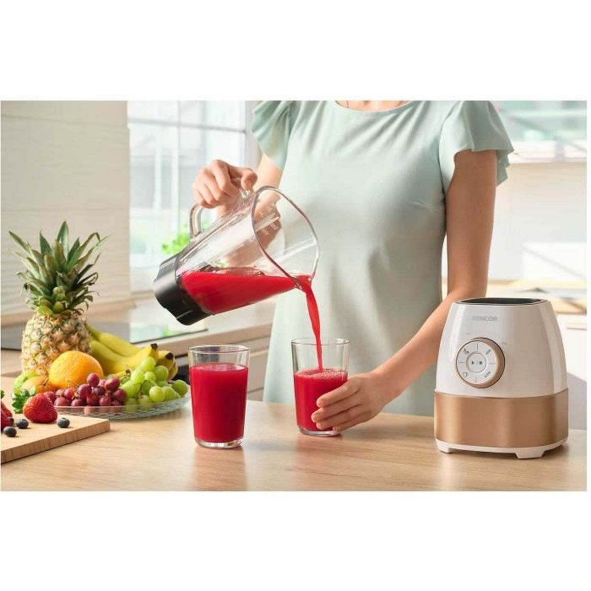 Eine Person gießt roten Saft aus einer Glaskanne in zwei Gläser neben Obst.