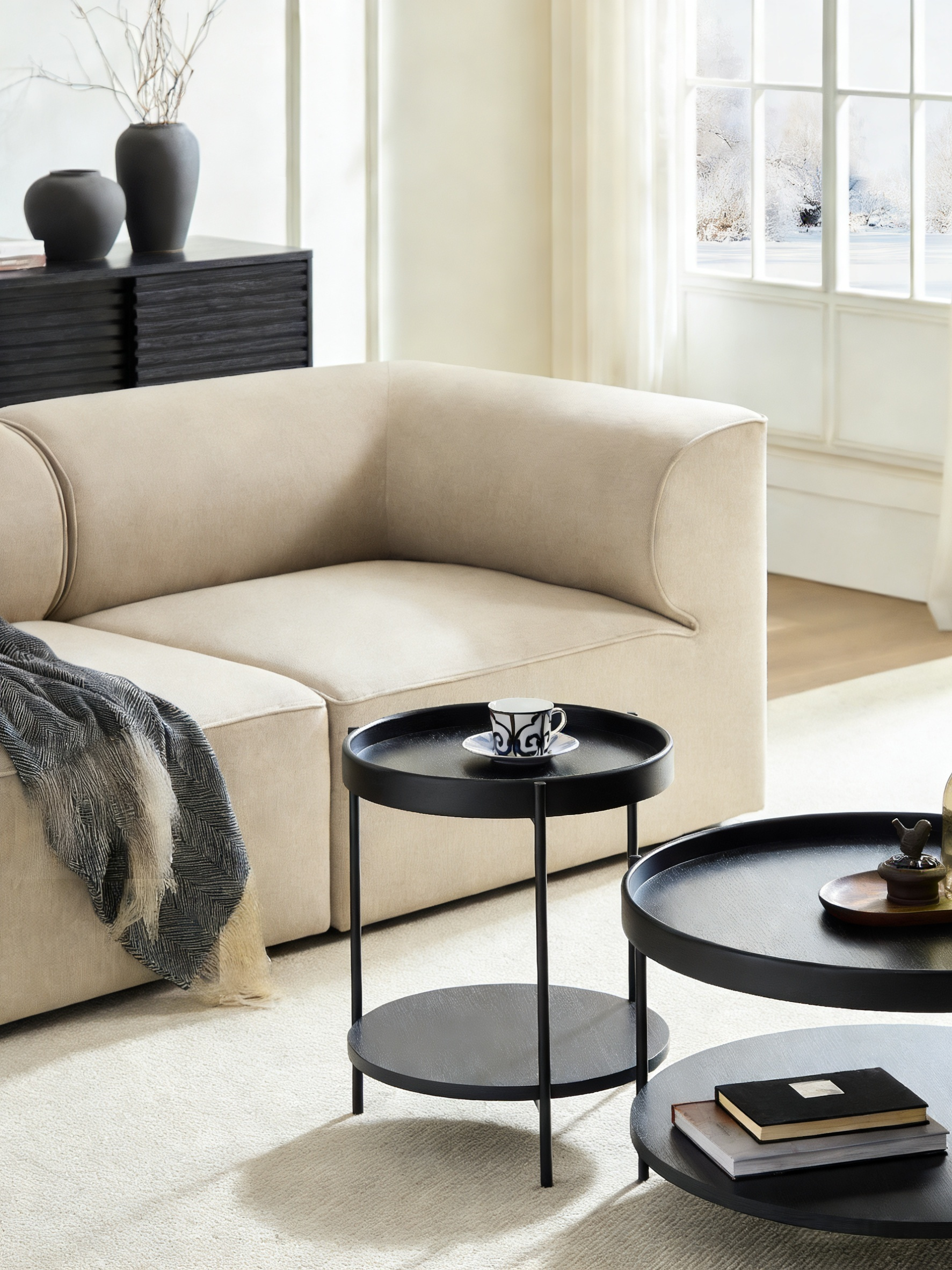 Ein beiges Sofa mit einer Decke, daneben schwarze Beistelltische mit Tasse und Büchern.