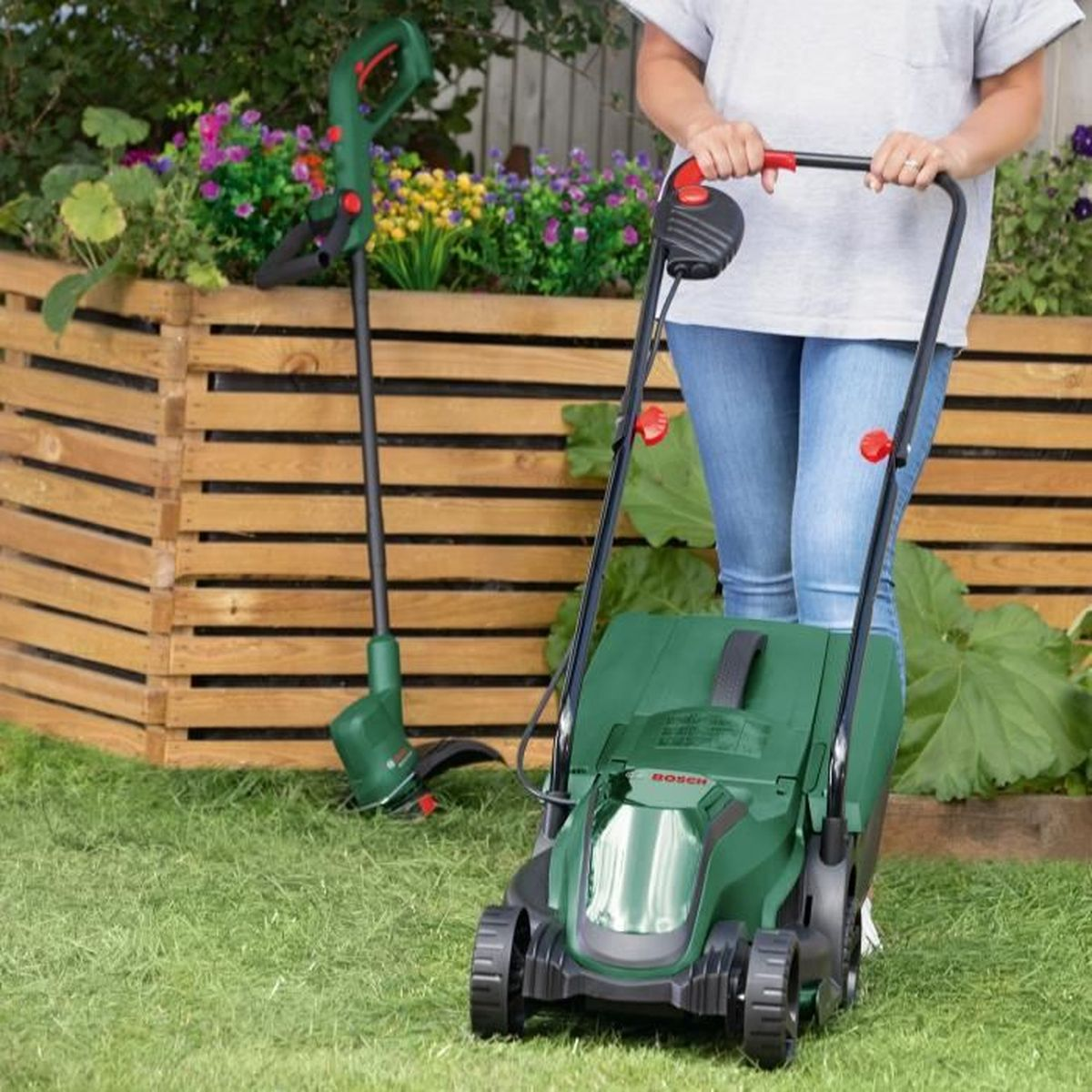 Eine Frau steht mit einem Rasenmäher und einem Trimmer im Garten.