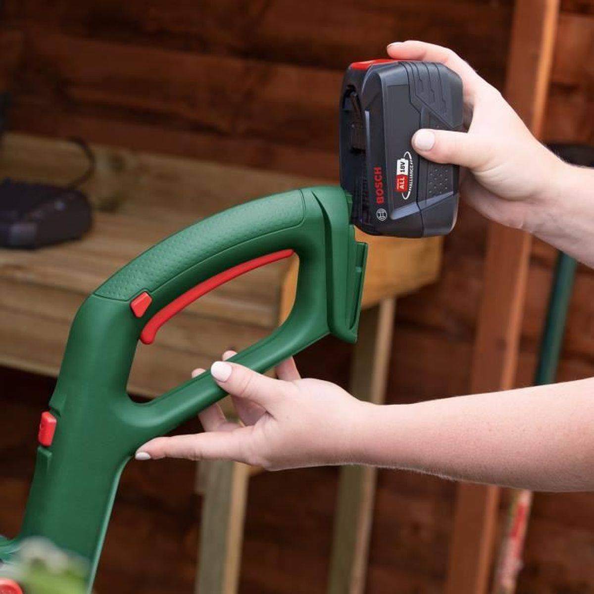 Hände stecken einen schwarzen Bosch 18V Akku in einen grünen Gartentrimmergriff.