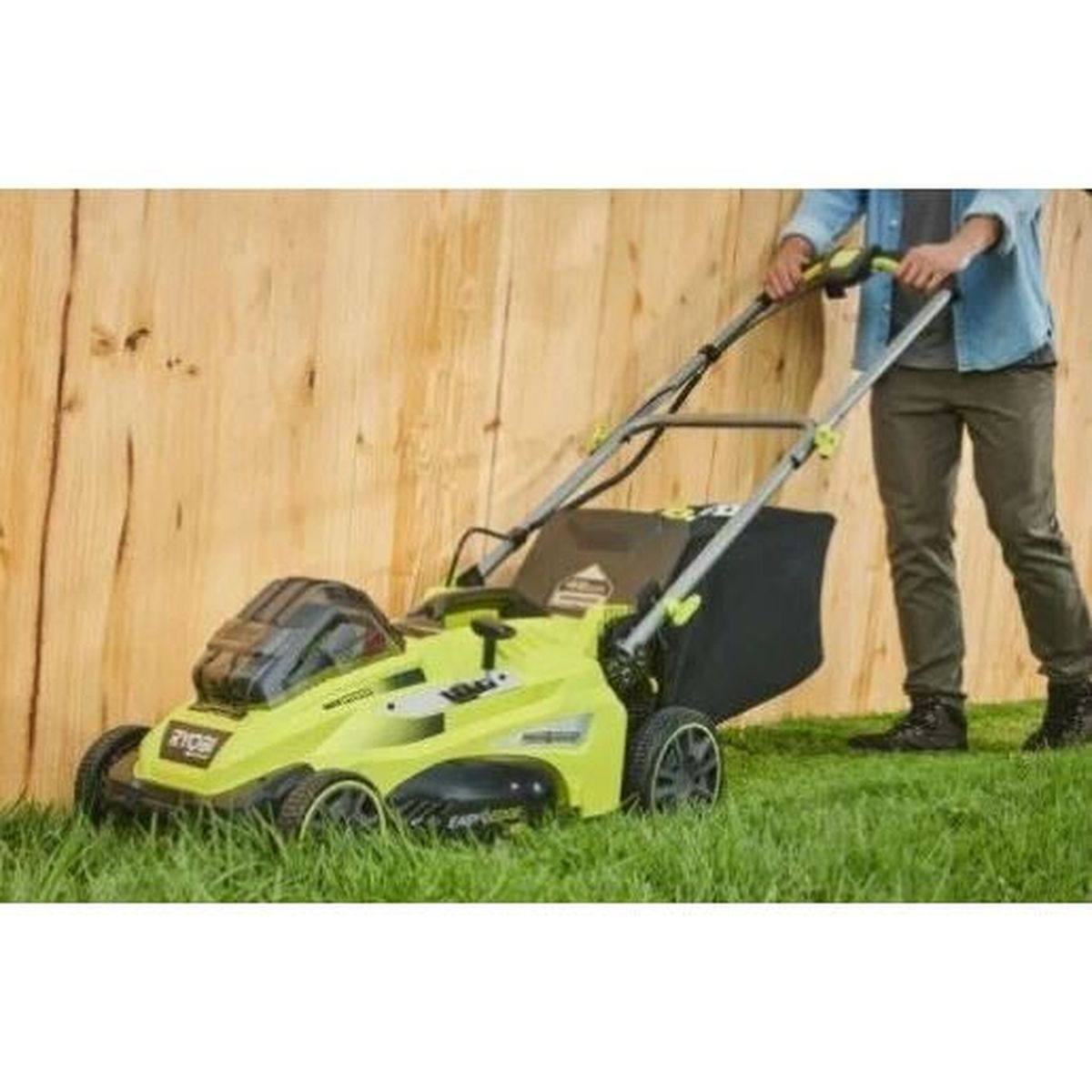 Ein Mann schiebt einen hellgrünen Rasenmäher auf Gras vor einer Holzwand.