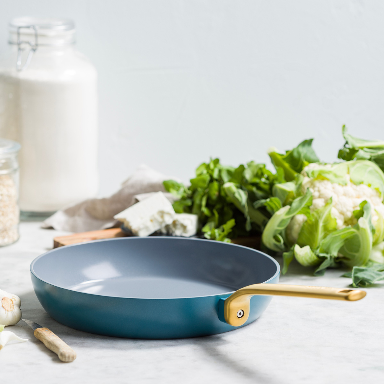 Eine blaue Bratpfanne mit goldenem Griff steht auf einer Marmorplatte. Zutaten wie Blumenkohl, Grünzeug, Käse und Knoblauch sind in der Nähe.