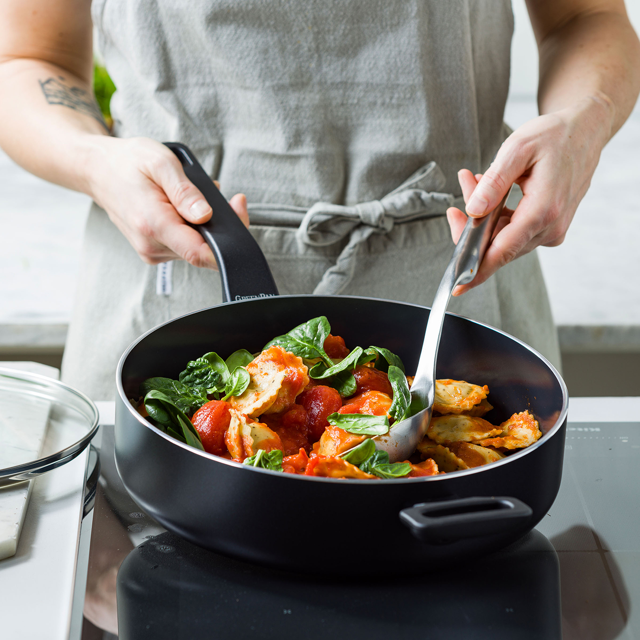 Eine Person in einer Schürze kocht Ravioli mit Tomaten und Spinat in einer Pfanne.