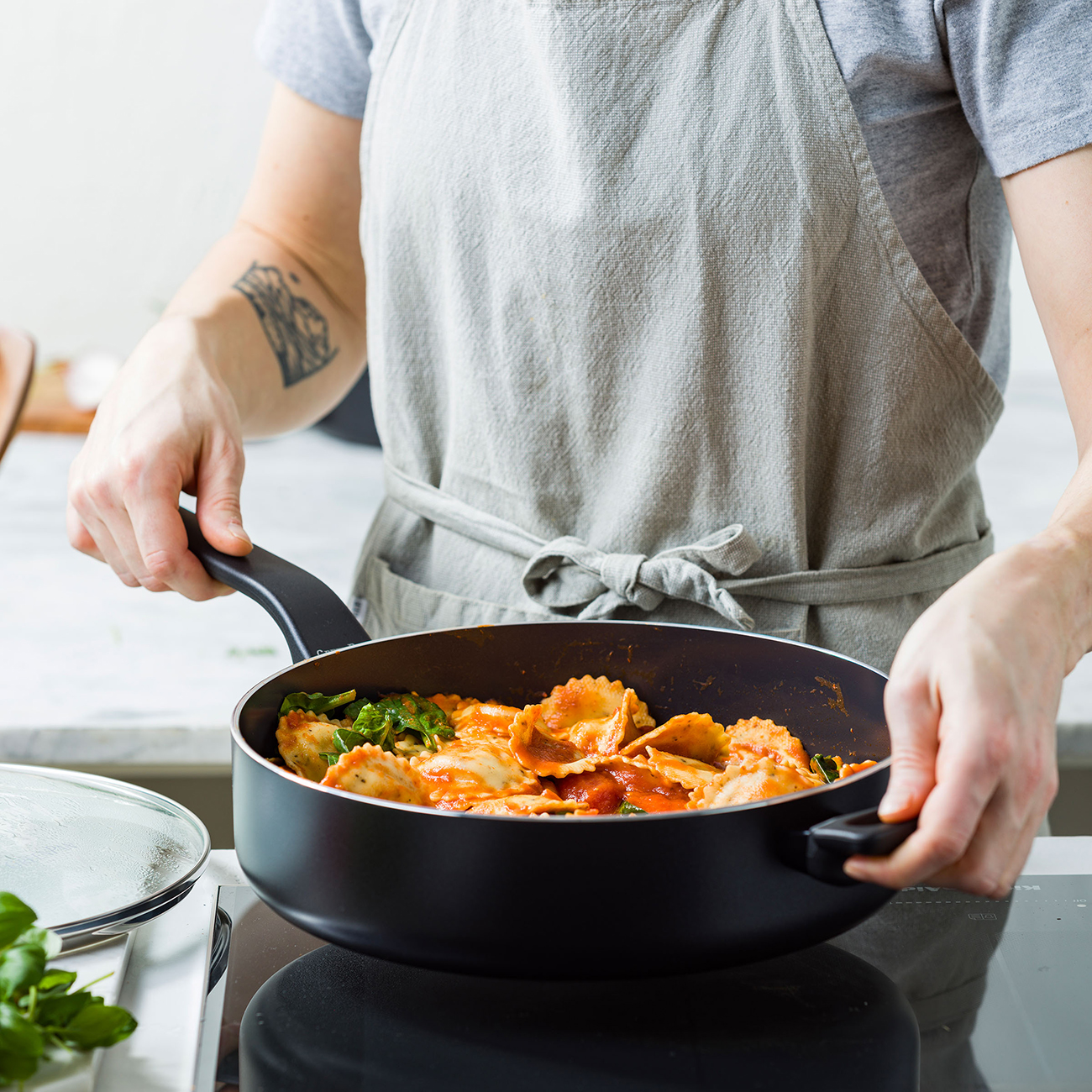 Ravioli in einer Pfanne mit Tomatensauce und Spinat, von einer Person mit Schürze gehalten.