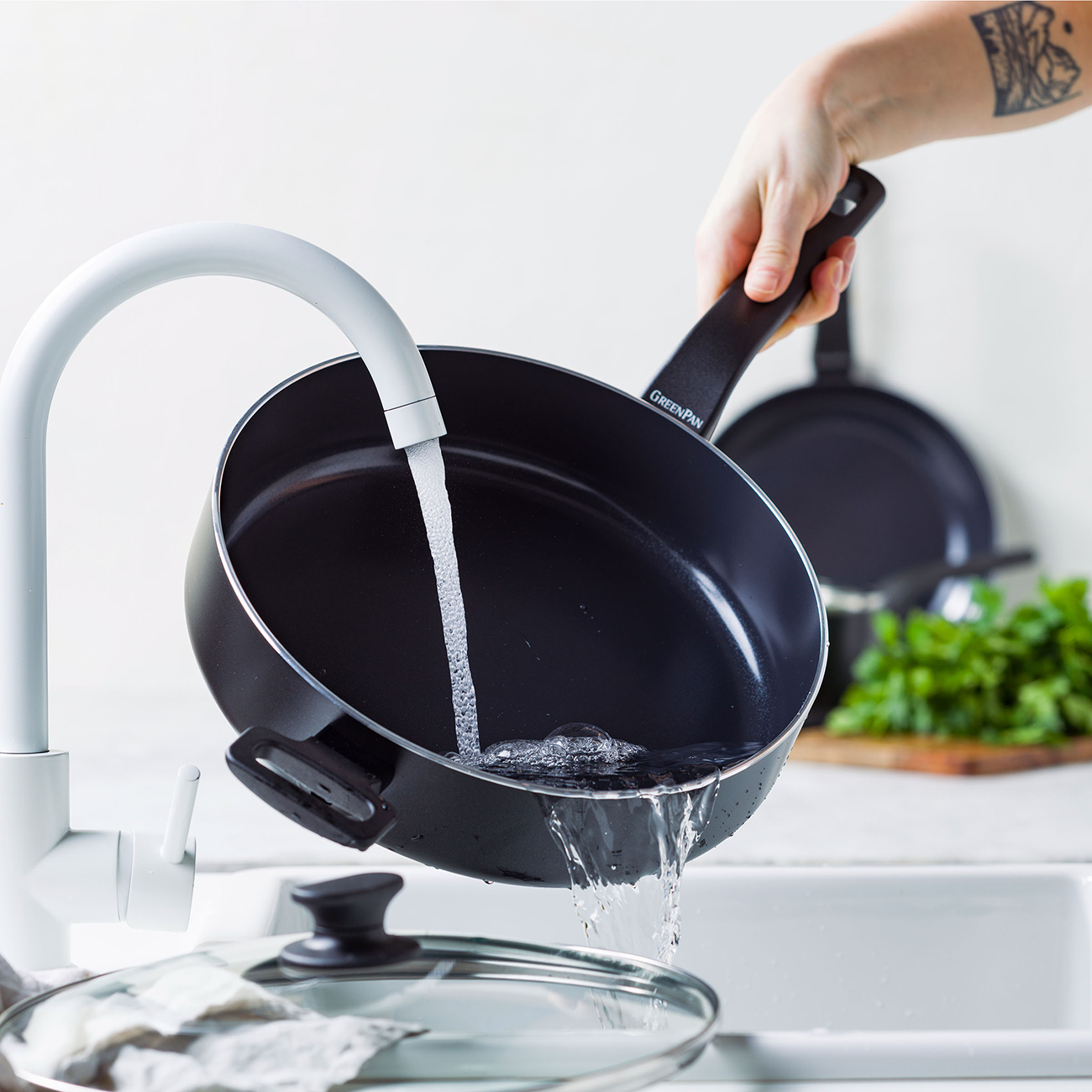 Eine Hand kippt eine schwarze Bratpfanne unter einem weißen Wasserhahn, Wasser fließt hinein.