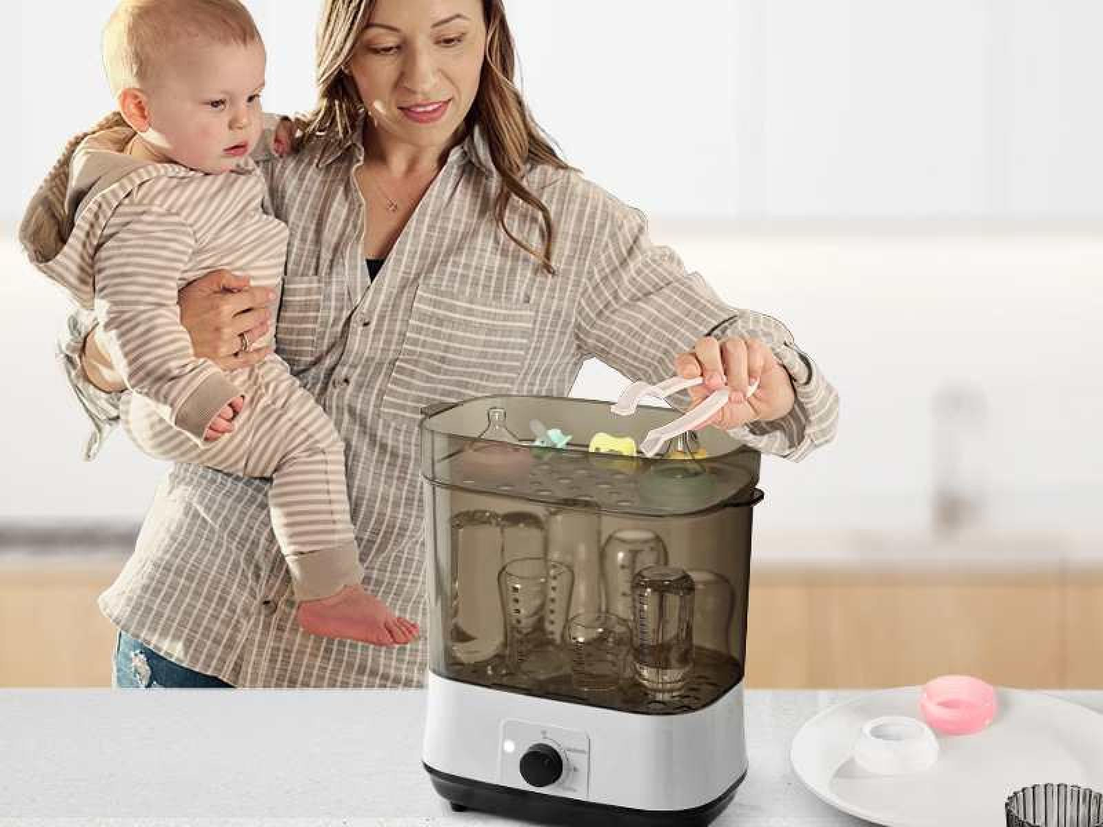 Frau hält Baby in der Nähe eines Flaschensterilisators mit Flaschen darin. Sie benutzt eine Zange.
