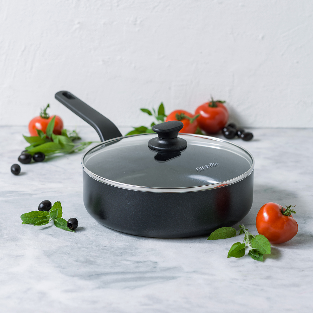 Schwarzer Kochtopf mit Glasdeckel, garniert mit Tomaten, Oliven und Kräutern.