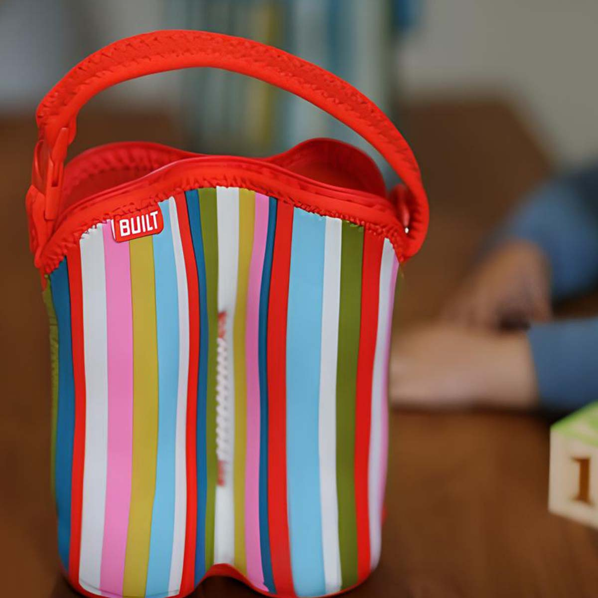Eine bunte, gestreifte Tasche mit rotem Griff und Reißverschluss steht auf einem Holztisch.