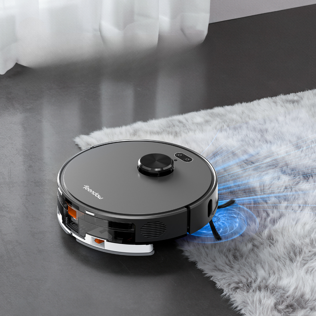 Un robot aspirapolvere grigio scuro pulisce un tappeto bianco e soffice su un pavimento grigio.