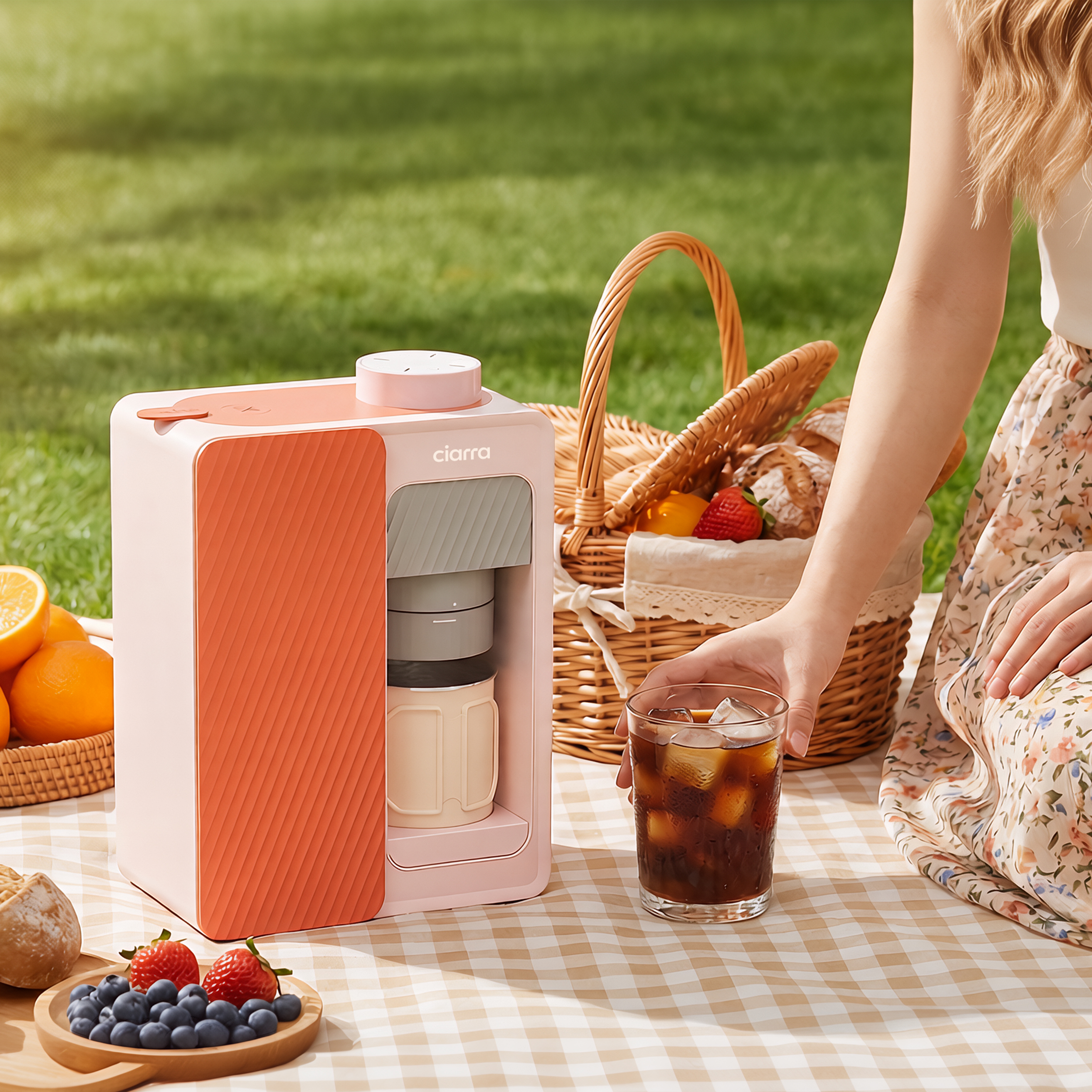 Ein rosa Kaffeevollautomat steht auf einer karierten Decke, daneben ein Picknickkorb mit Obst und Brot.
