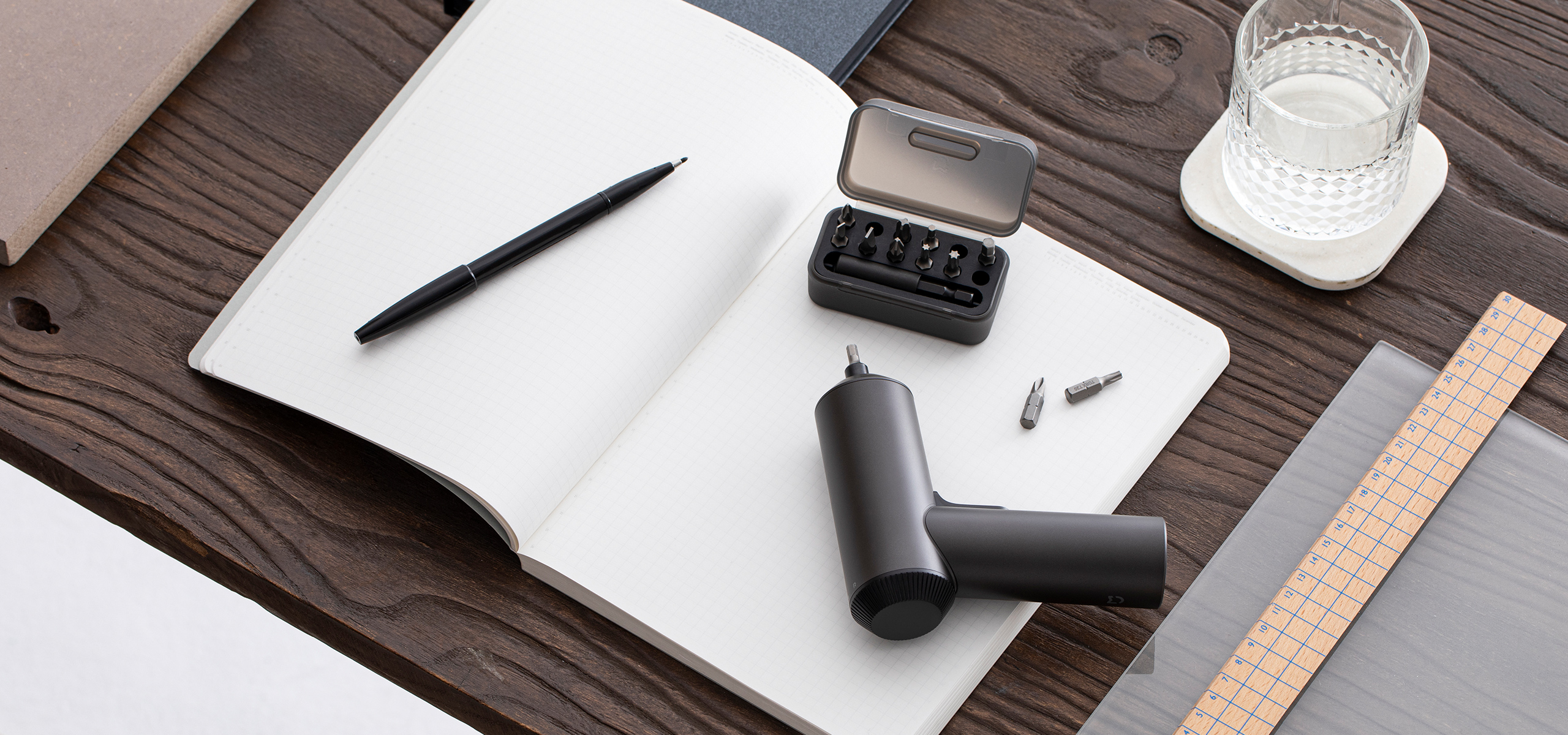 Tournevis noir avec embouts, carnet, stylo et verre d'eau sur table en bois.