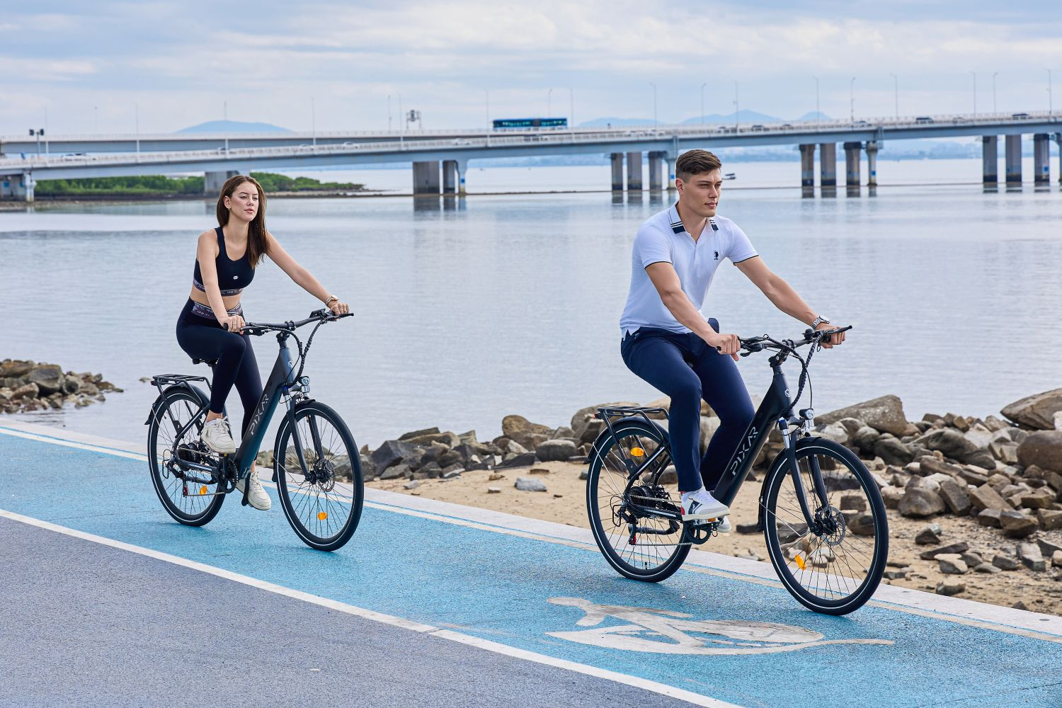 Hombre y mujer en bicicleta por sendero junto al agua, puente al fondo.