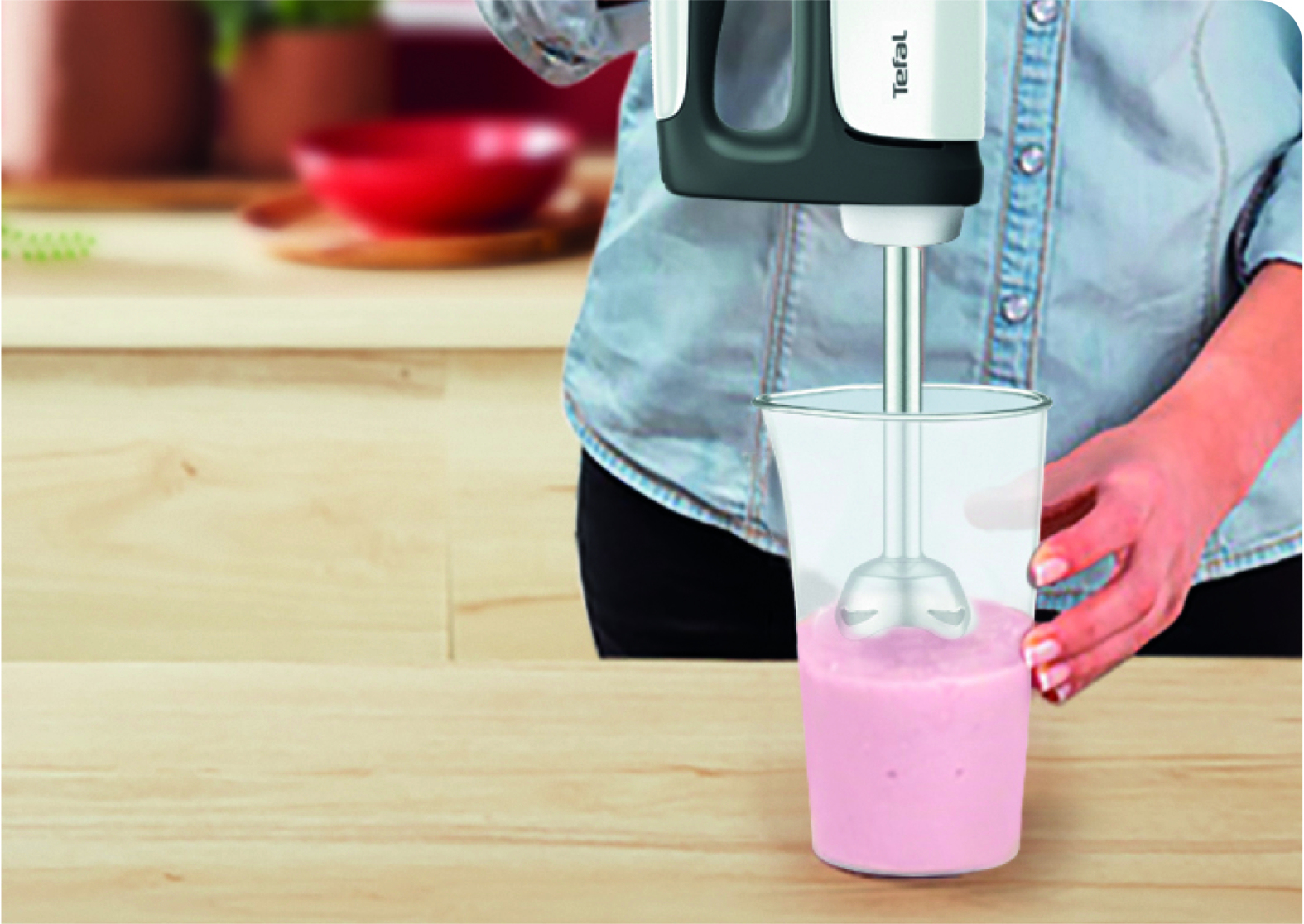 A hand blender mixes a pink smoothie in a glass on a wooden table.