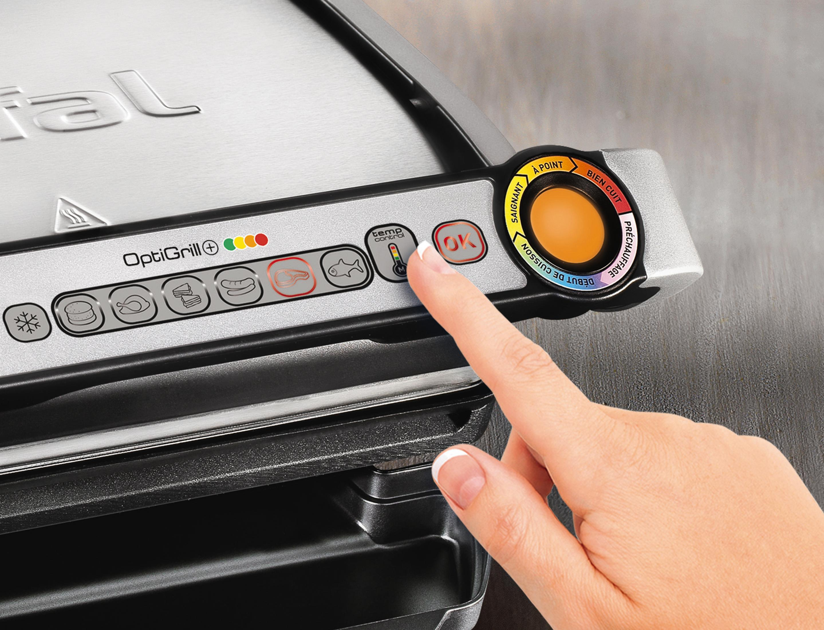 Una mano preme il pulsante OK su un Tefal OptiGrill argentato con programmi di cottura.