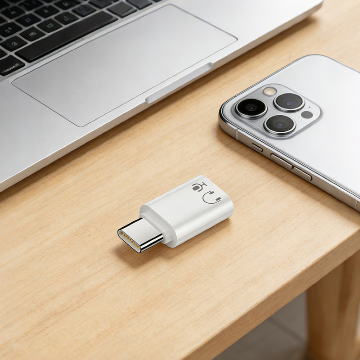 Biały adapter USB-C z ikonami słuchawek i mikrofonu leży na drewnianym biurku obok laptopa i telefonu.