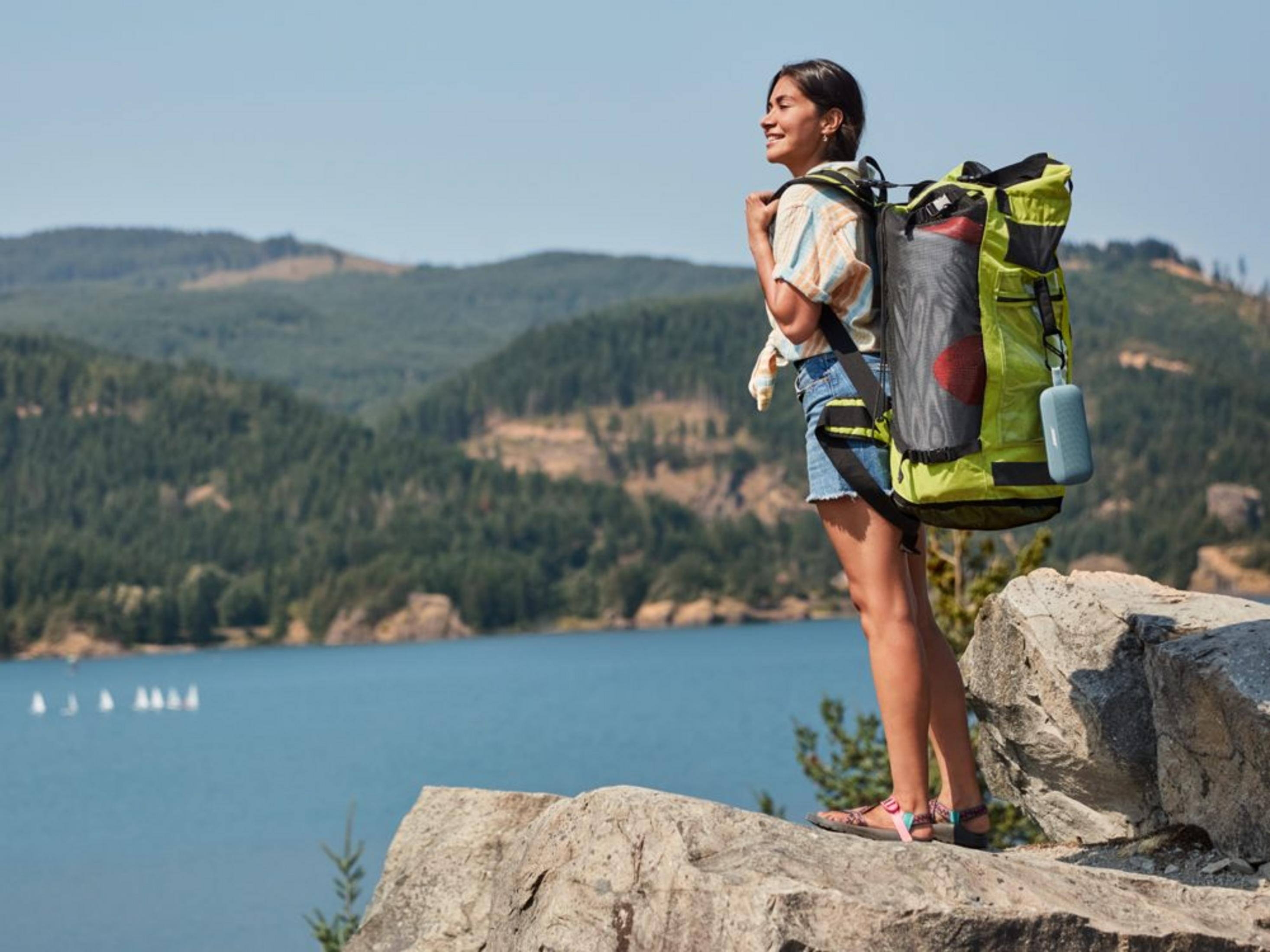 Mujer con mochila sobre rocas junto al lago con veleros y colinas boscosas detrás.